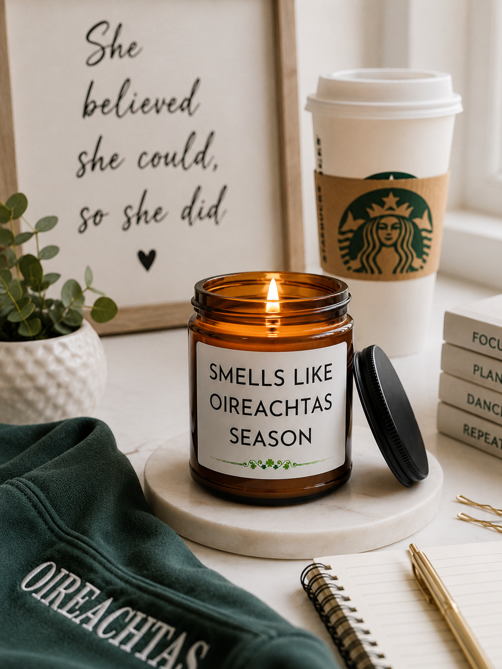 An amber glass jar candle with a black lid and a white label reading 'SMELLS LIKE OIREACHTAS SEASON' sits on a white marble coaster in a styled desk scene. A Starbucks coffee cup, framed quote print, plant, notebook, pen, and a dark green sweatshirt are arranged around it.