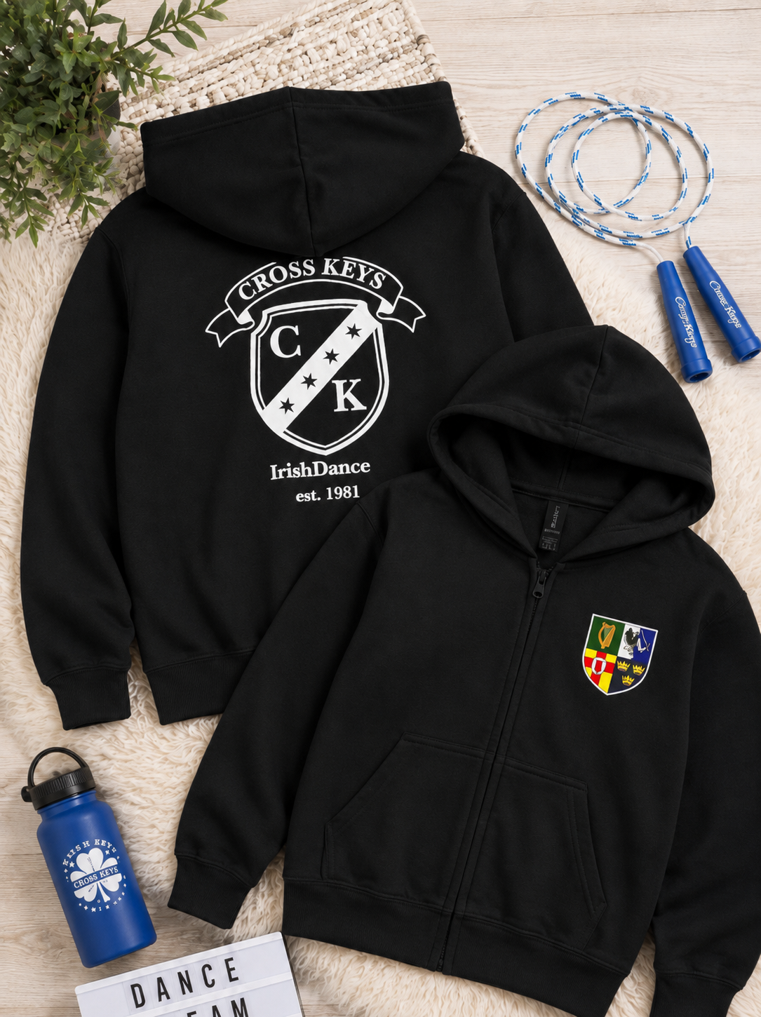 A black zip-up hoodie and a black pullover hoodie are arranged on a beige textured rug with lifestyle props, including a blue water bottle, jump rope, and a plant. The back of the pullover hoodie features a large white 'Cross Keys Irish Dance' crest graphic, and the front hoodie has a smaller matching crest on the left chest.
