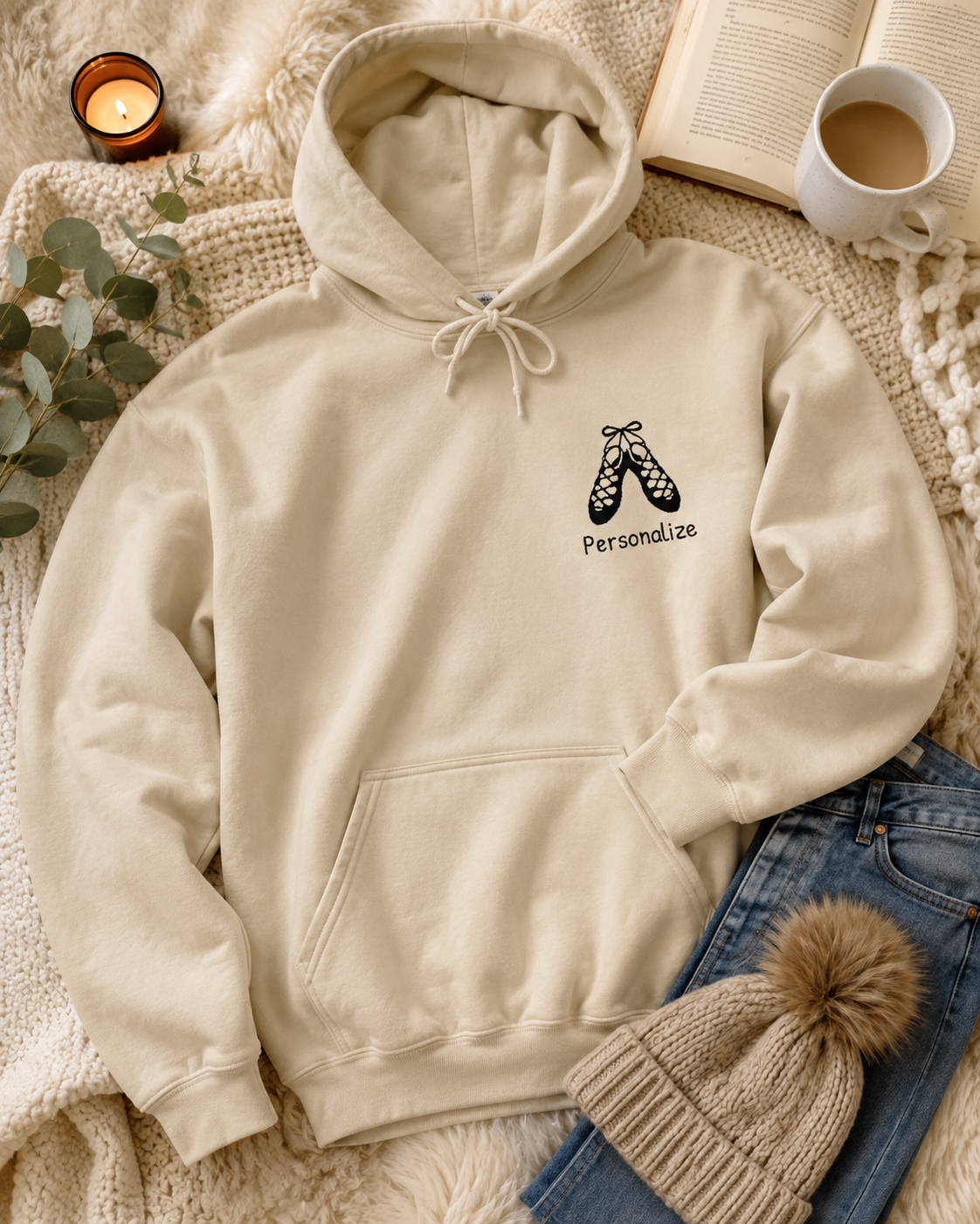 A cream-colored hoodie with a black graphic of ballet shoes and the word 'Personalize' on the left chest, styled in a cozy flat-lay with a book, a mug of coffee, a candle, eucalyptus leaves, blue jeans, and a knit beanie.