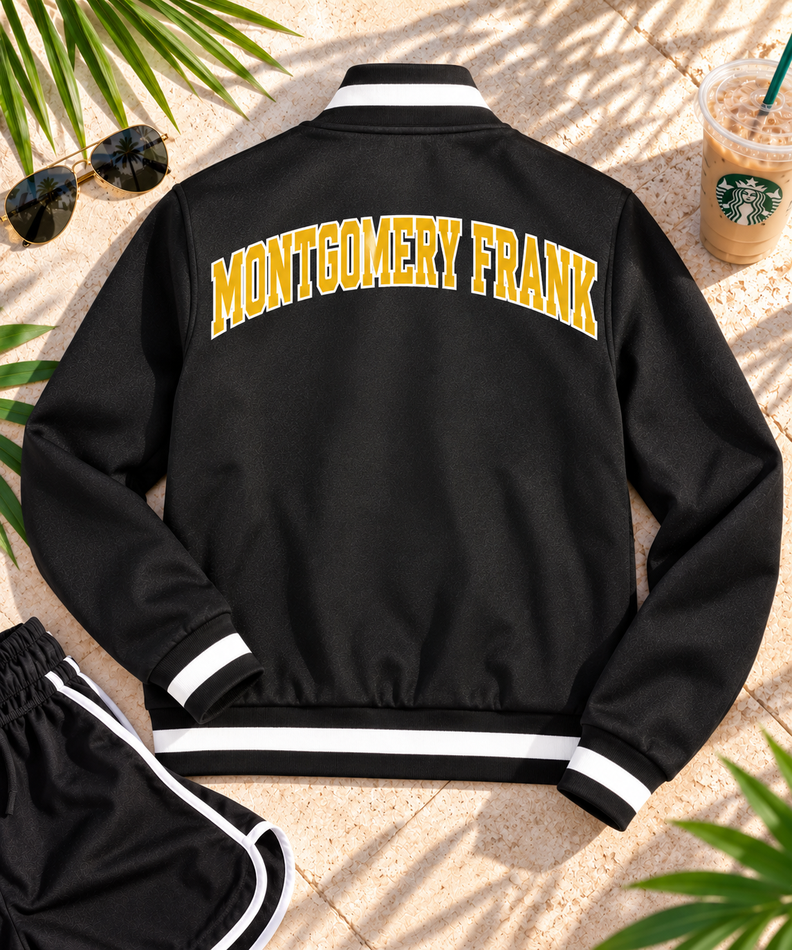 A black varsity-style jacket is laid flat on a stone surface, shown from the back with large yellow 'MONTGOMERY FRANK' lettering across the upper back and white striped ribbed collar, cuffs, and hem. Sunglasses, palm leaves, and a Starbucks iced coffee are visible around it as styled props.