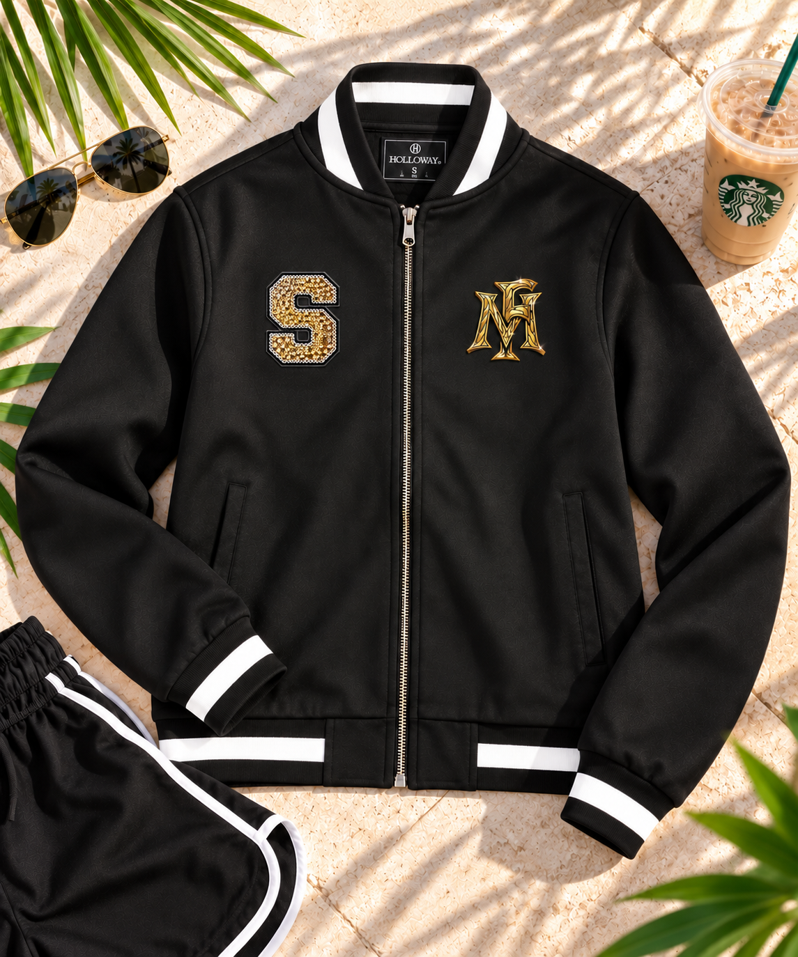 A black varsity-style bomber jacket laid flat on a sandy surface with palm leaves and a Starbucks cup nearby. The jacket has white striped ribbed collar, cuffs, and hem, a front zipper, and two gold embroidered chest graphics: a sequin 'S' and a gold 'M'.