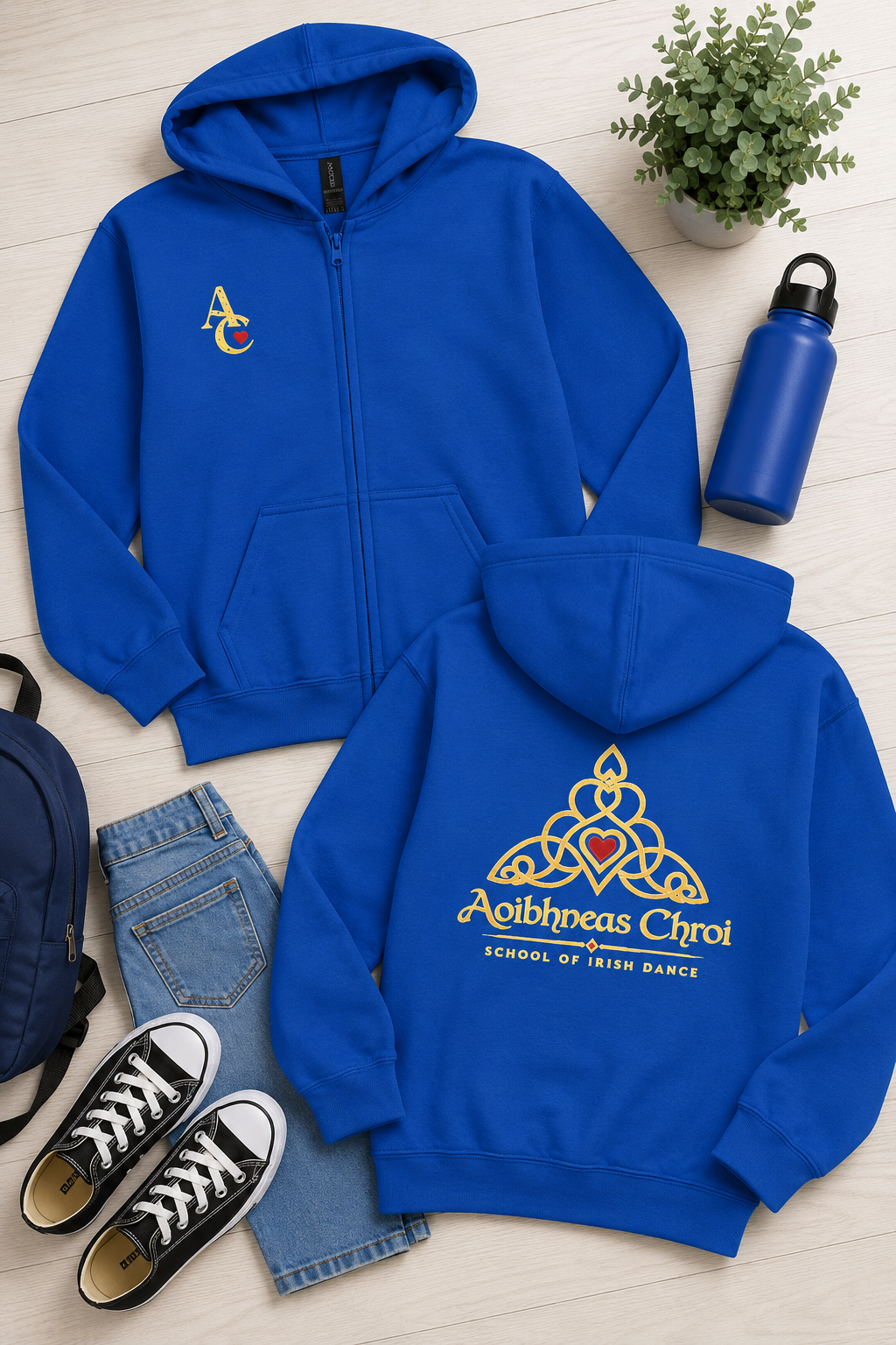 Two royal blue zip-up hoodies are arranged on a light wood floor with jeans, black sneakers, a blue water bottle, a backpack, and a small potted plant. The front hoodie has a small embroidered multicolor logo on the left chest, and the back hoodie has a large gold Celtic-style crest with a red heart and the text 'Aoibhneas Chroi' and 'School of Irish Dance'.