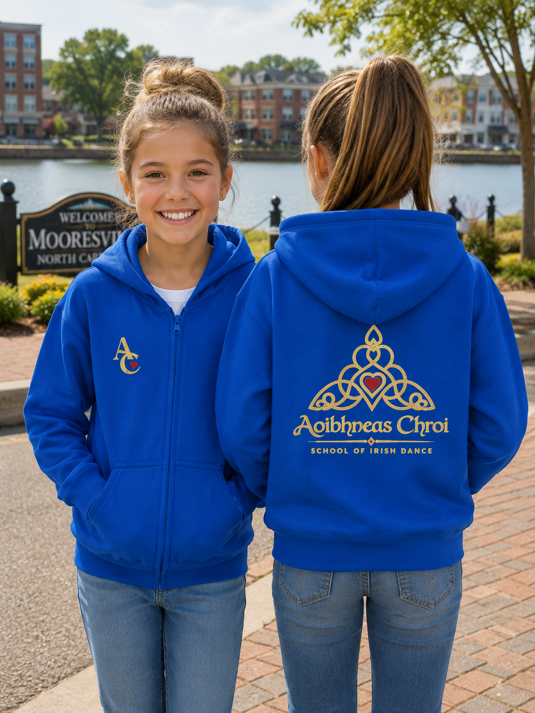Two girls modeling a royal blue zip-up hoodie outdoors near a waterfront. The girl on the left shows the front with a small gold and red logo on the chest, and the girl on the right shows the back with a large gold Celtic knot design and the text 'Aoibhneas Chroi Schools of Irish Dance'.