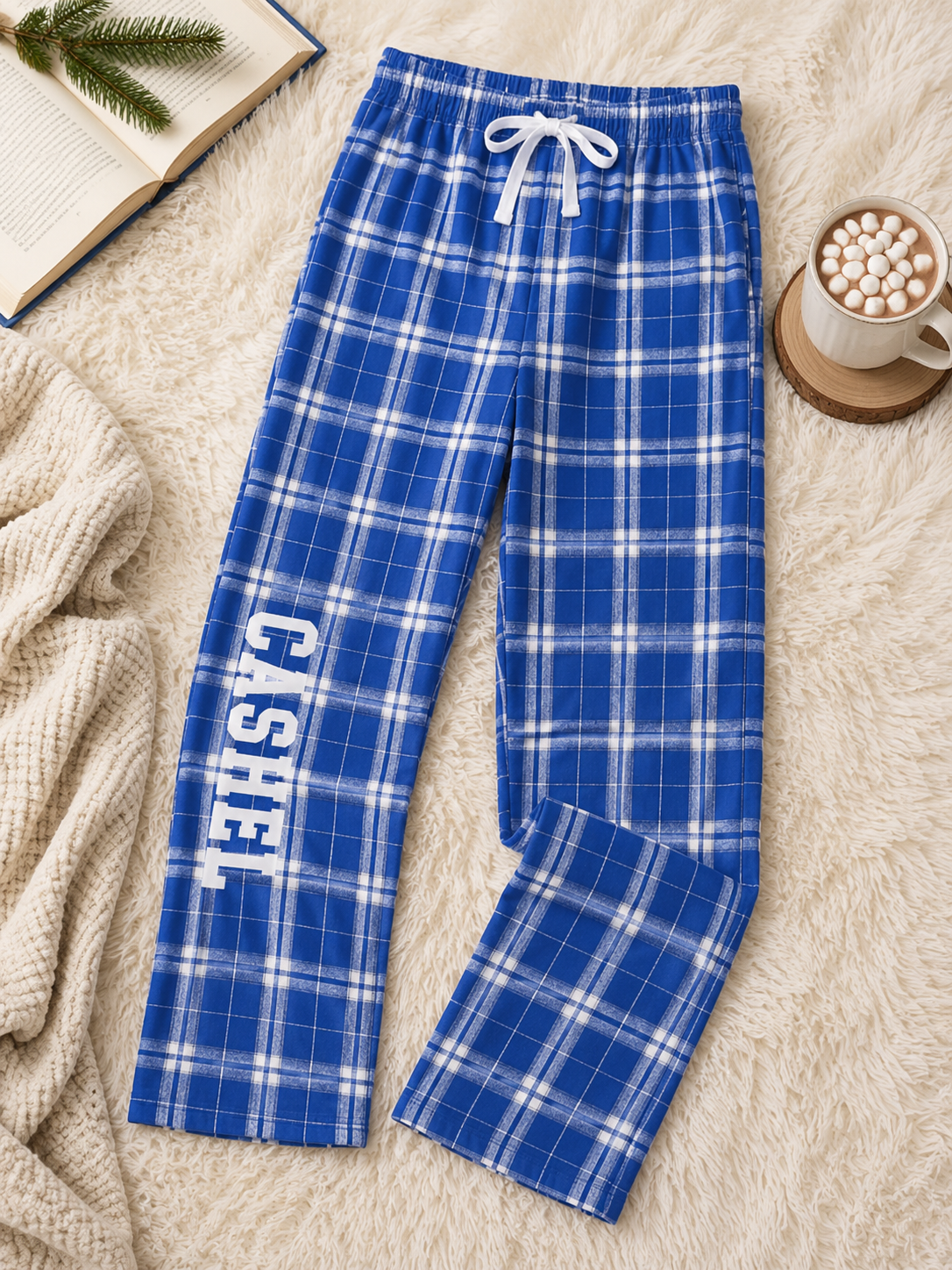 Blue and white plaid pajama pants with 'CASHEL' printed vertically on one leg, styled in a cozy flat-lay arrangement on a white faux fur rug with a mug of hot chocolate, an open book, and a knit blanket.