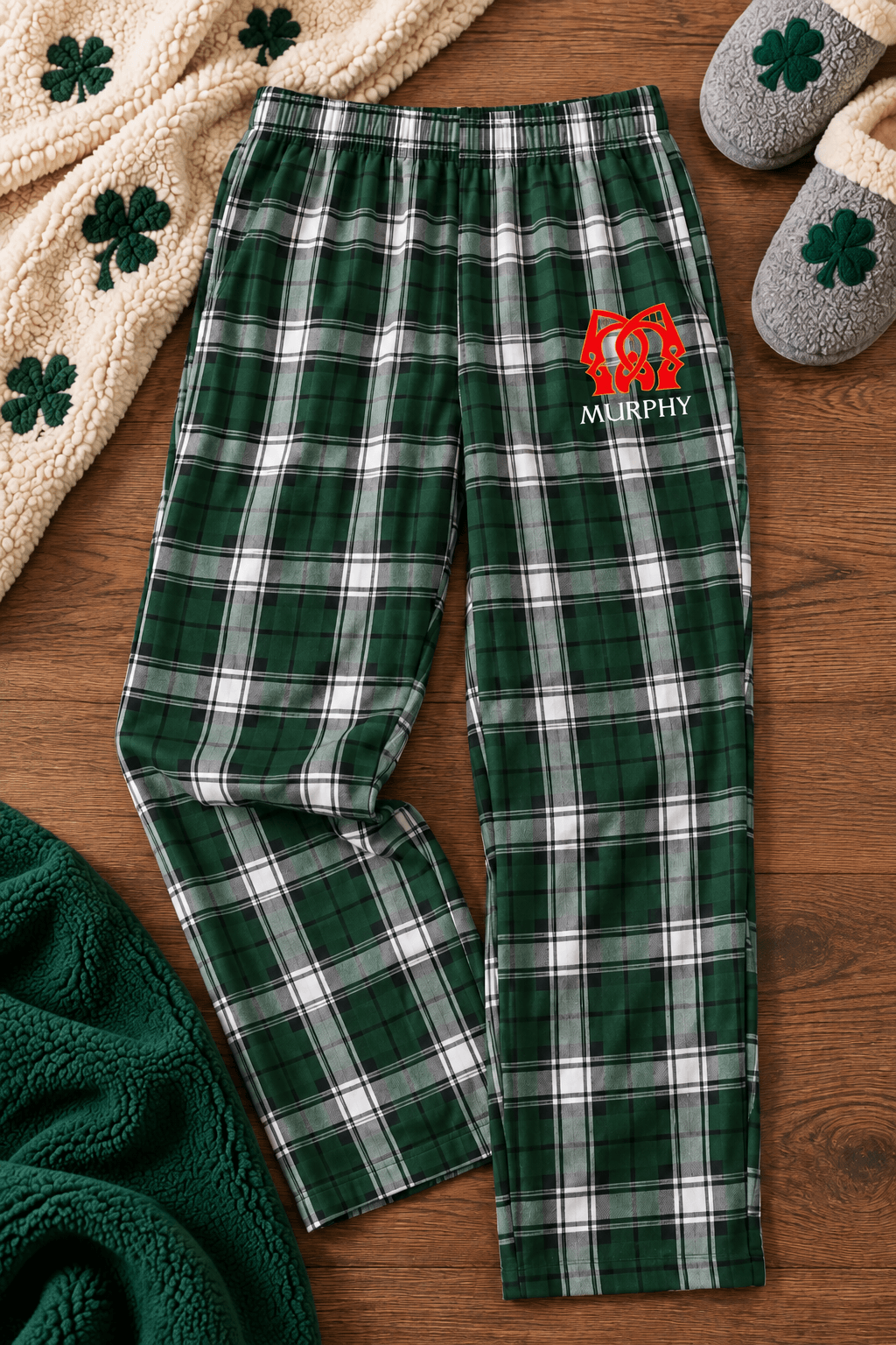 A pair of green and white plaid pajama pants with a red embroidered monogram and the name 'MURPHY' on the left thigh, laid flat on a wooden surface with cozy holiday-themed props including slippers, a blanket, and a folded green knit blanket.