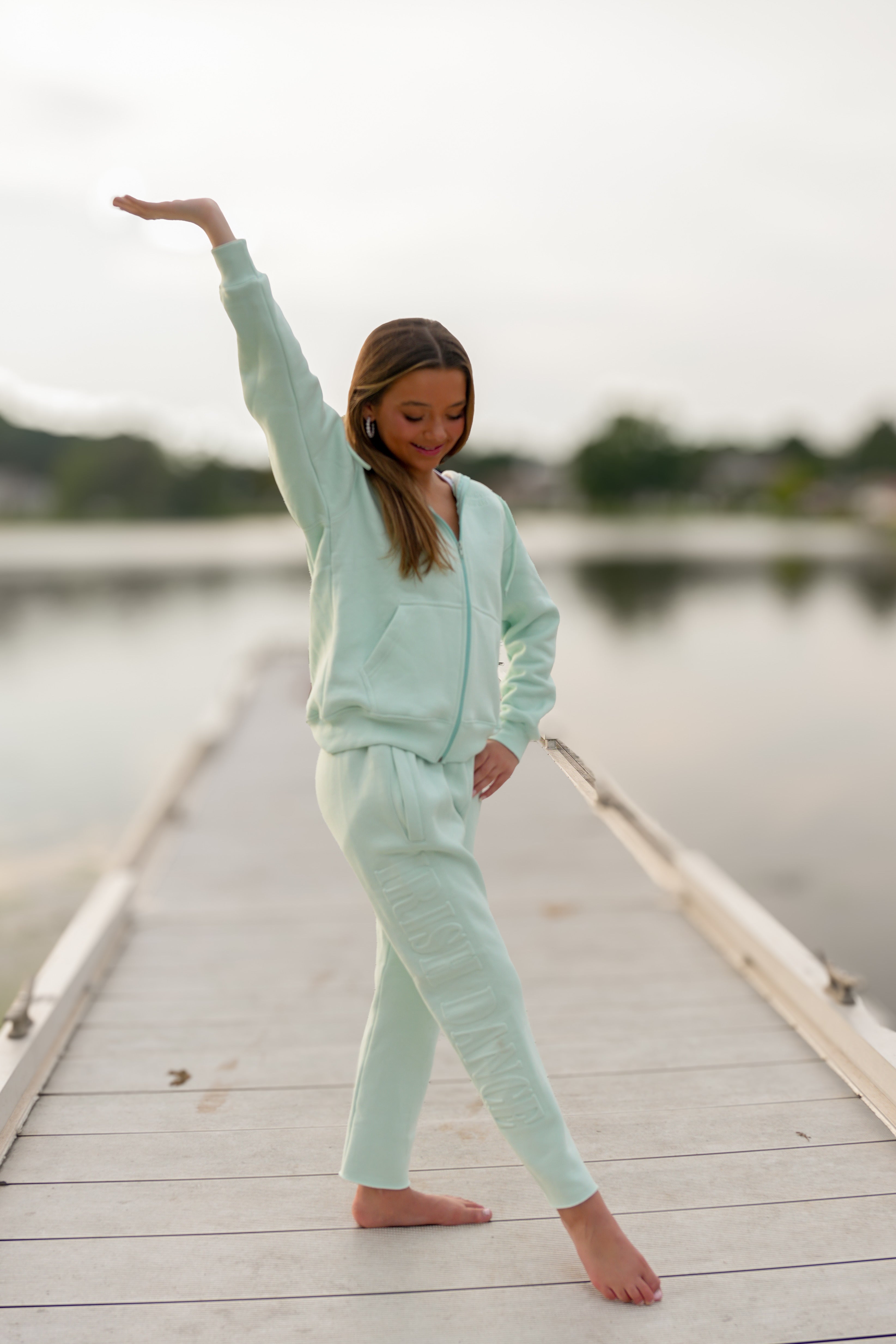 A young girl in a light mint green two-piece lounge set poses on a wooden dock over calm water, stretching her arm overhead.