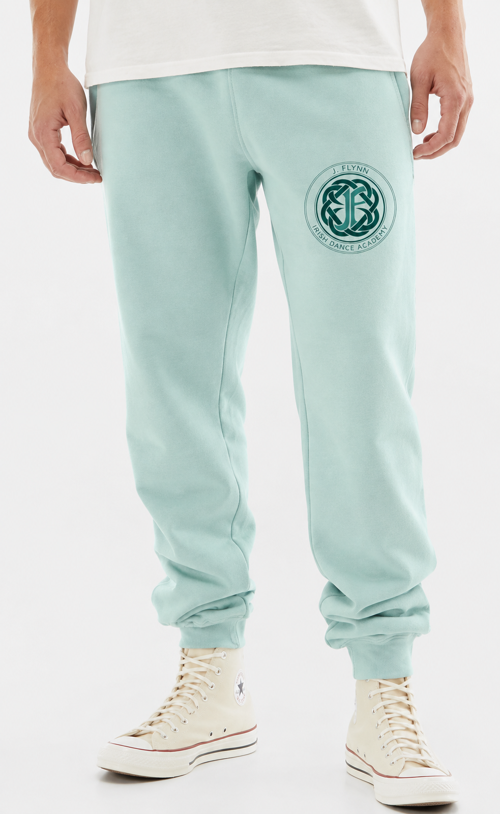 A person wearing mint green sweatpants with a circular Celtic knot logo on the left thigh, paired with a white t-shirt and off-white high-top sneakers, shown from the waist down against a white studio background.