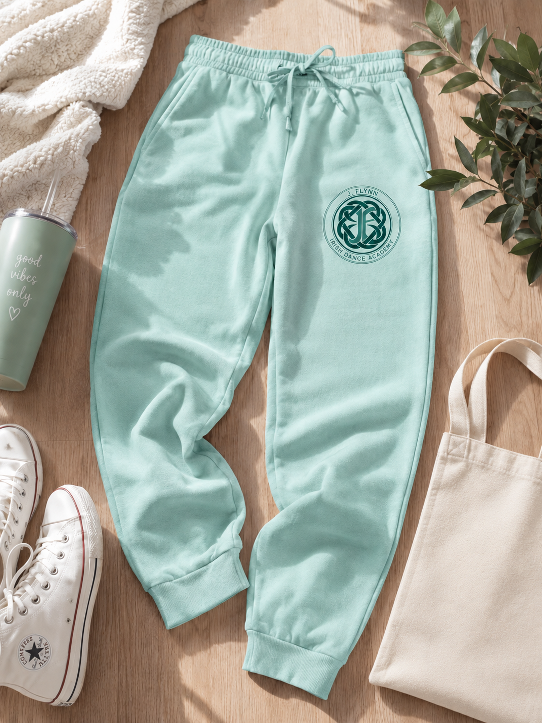 A pair of light mint green jogger sweatpants with a dark green circular Celtic knot logo on the left thigh, styled in a flat lay on a light wood floor with a white tumbler, white sneakers, a canvas tote bag, and a cream knit blanket.