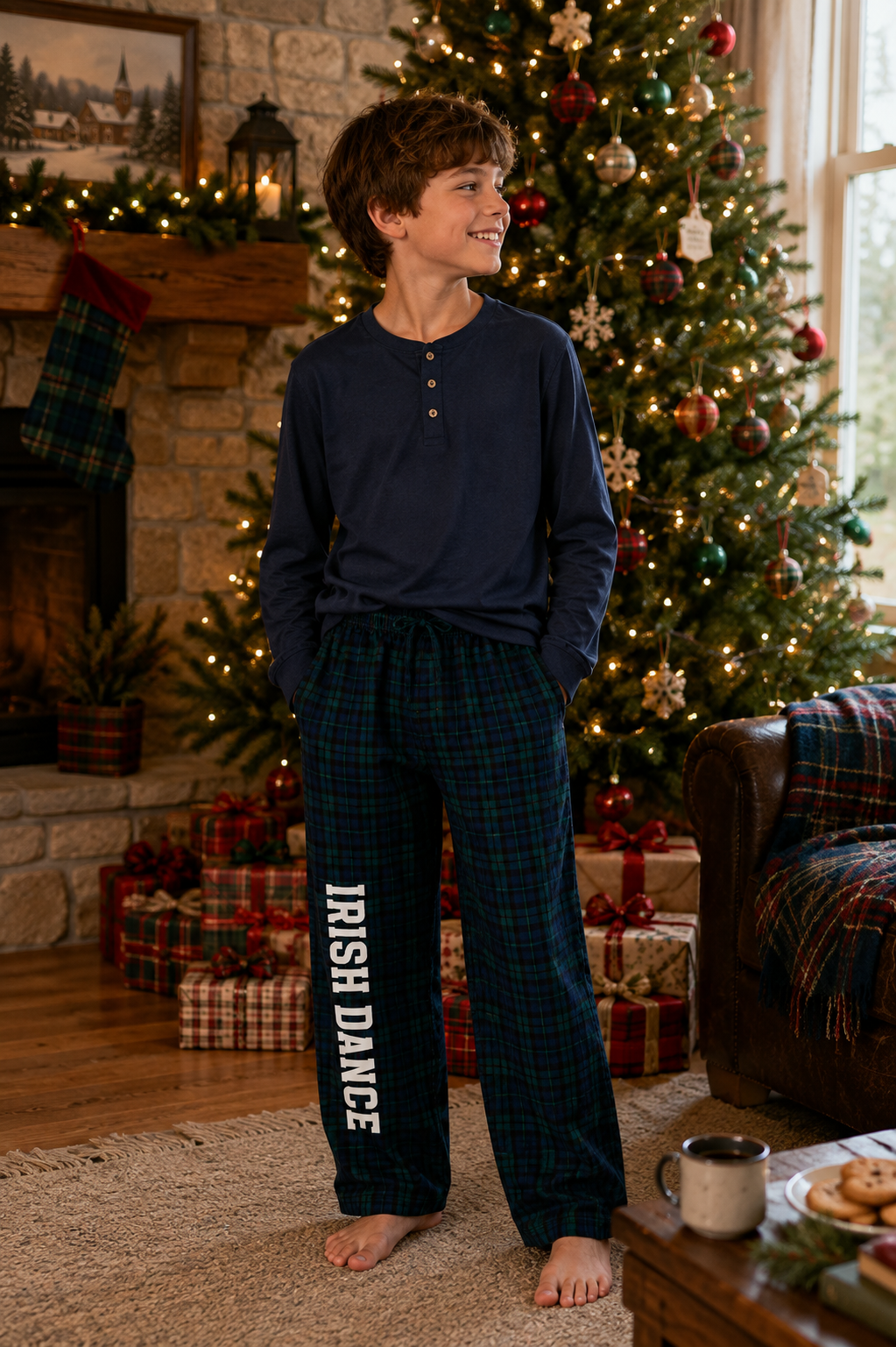 A boy stands barefoot in a cozy living room decorated for Christmas, wearing a navy long-sleeve henley shirt and dark green plaid pajama pants with 'IRISH DANCE' printed vertically in white on one leg.