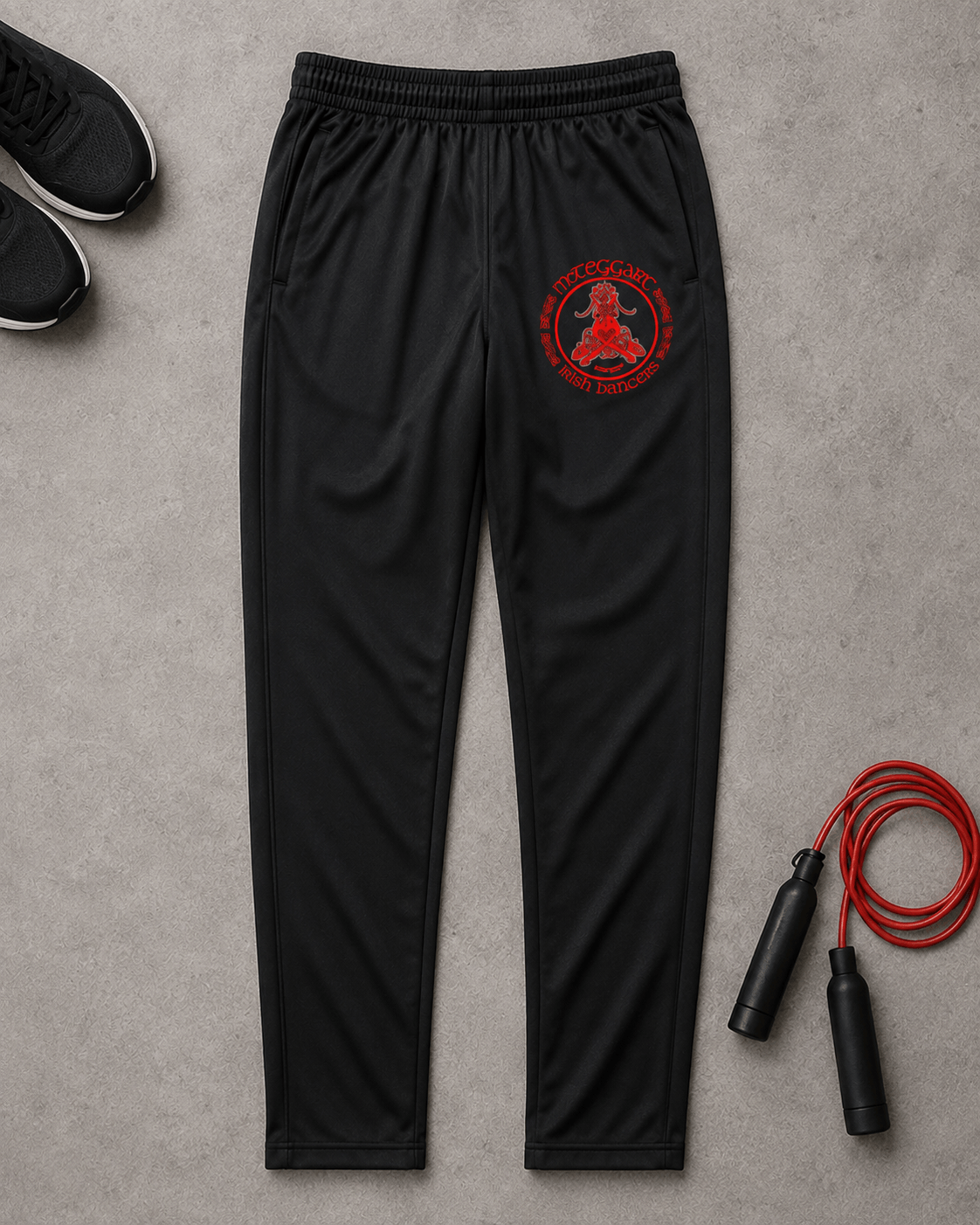Black athletic jogger pants with a red circular logo on the left thigh, laid flat on a gray concrete surface with sneakers, a jump rope, and a small towel as styling props.