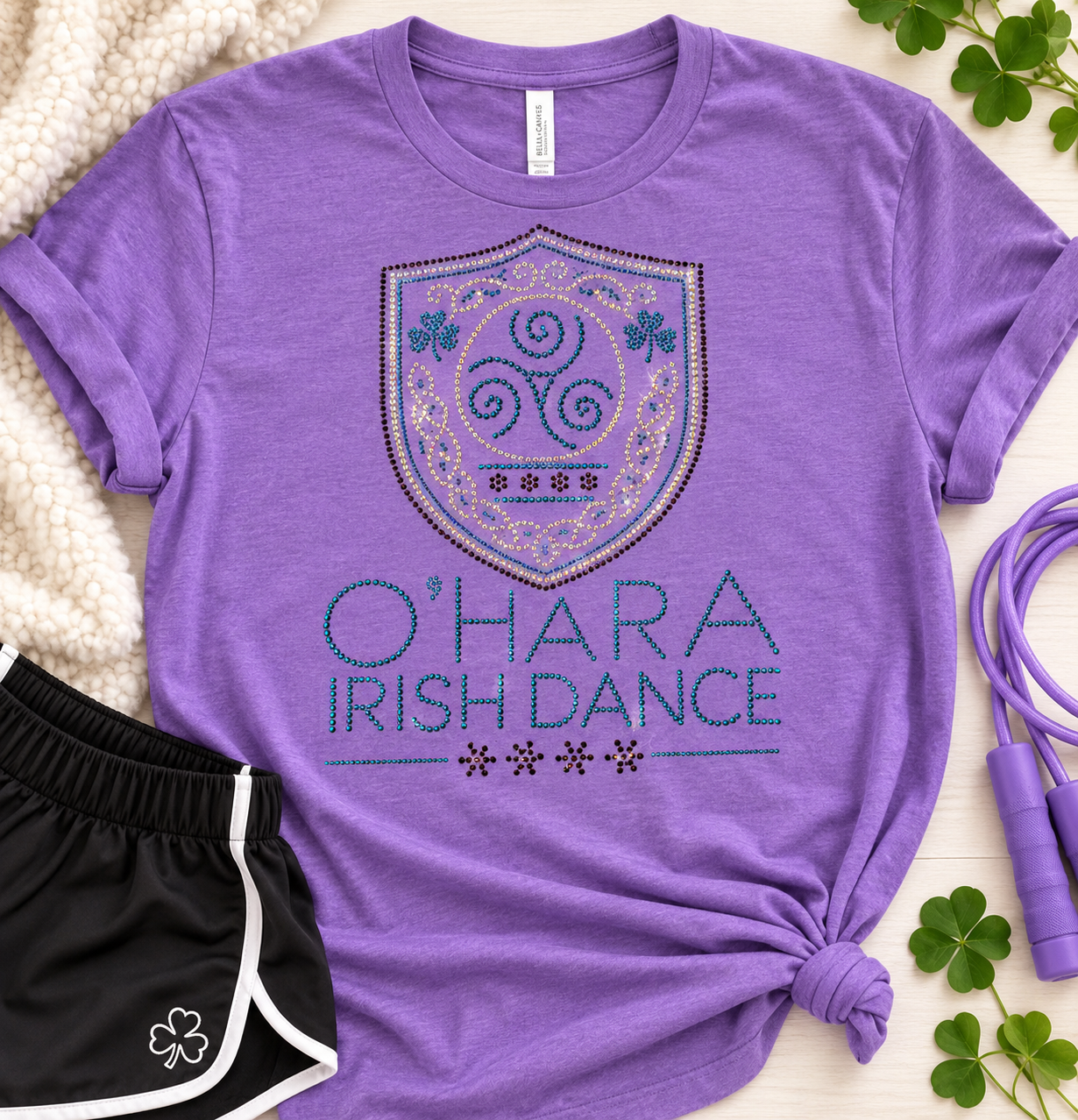 Flat lay of the heather purple O'Hara Irish Dance rhinestone tee with Celtic crest, shamrocks and aqua rhinestone lettering