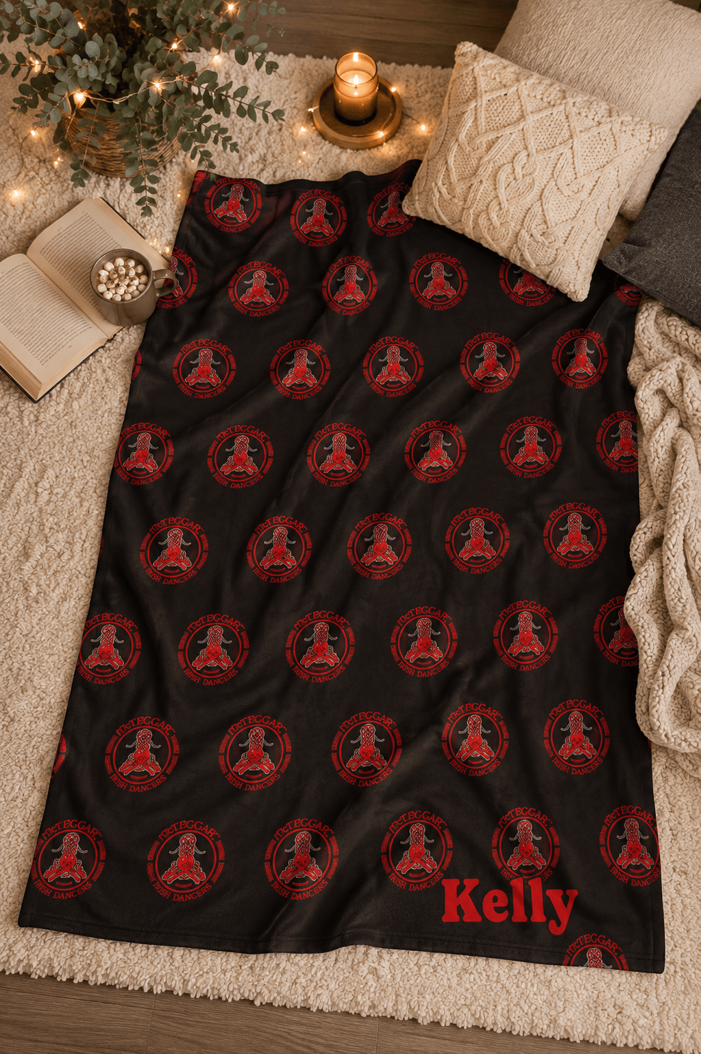 A black blanket or throw laid out on a cream rug, featuring a repeating red circular medallion pattern with a meditating figure and the name 'Kelly' in red at the bottom right. The scene is styled with pillows, a candle, books, and warm string lights.