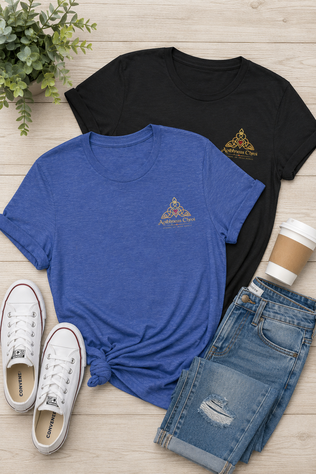 Two short-sleeve graphic T-shirts laid flat on a light wood surface, one black and one heather blue, both with a small gold triangular logo on the left chest. White sneakers, a coffee cup, and folded distressed jeans are styled around them as props.