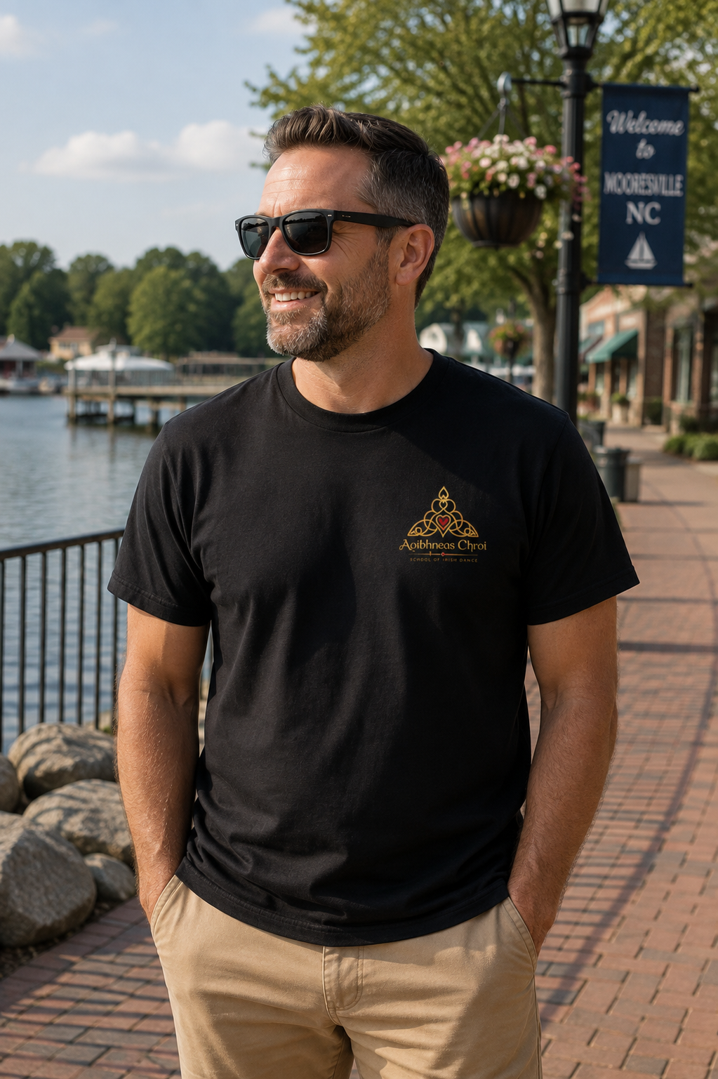 A man wearing a black t-shirt with a gold and red triangular graphic on the left chest, standing outdoors on a brick walkway near water with trees and buildings in the background.