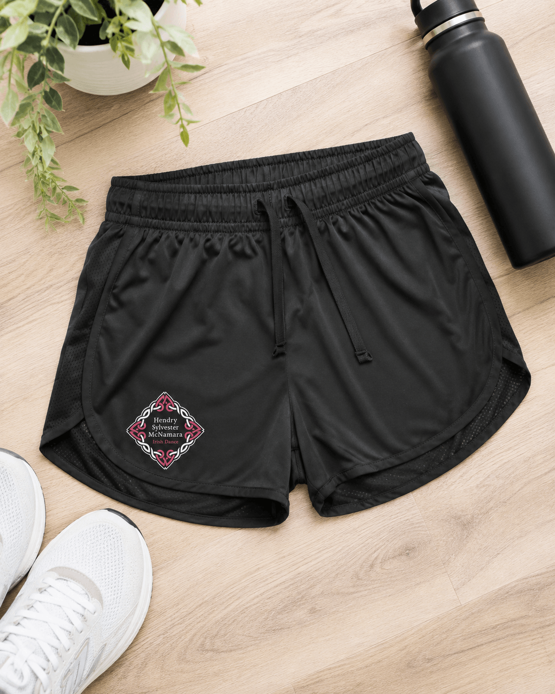 Black athletic shorts with a drawstring waist and a decorative embroidered logo patch on the left leg, laid flat on a light wood floor with white sneakers, a black water bottle, and a potted plant nearby.
