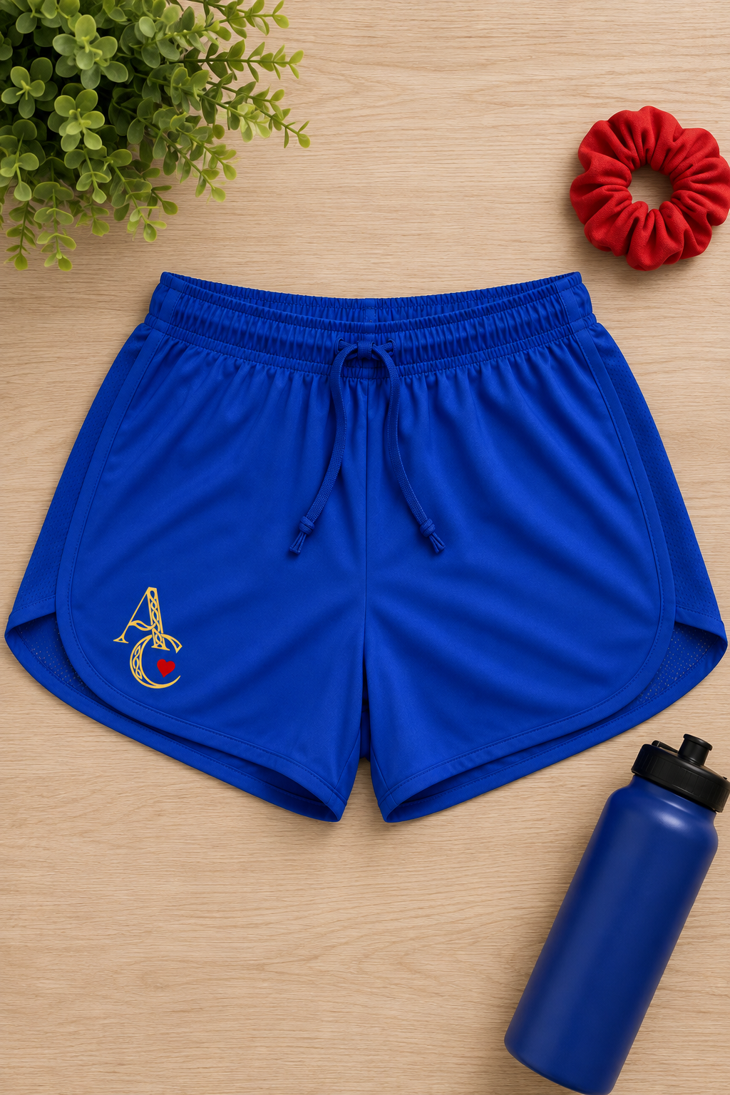 Royal blue athletic shorts with a drawstring waist and a small embroidered logo on the left leg, laid flat on a light wood background with a plant, red scrunchie, and blue water bottle as props.