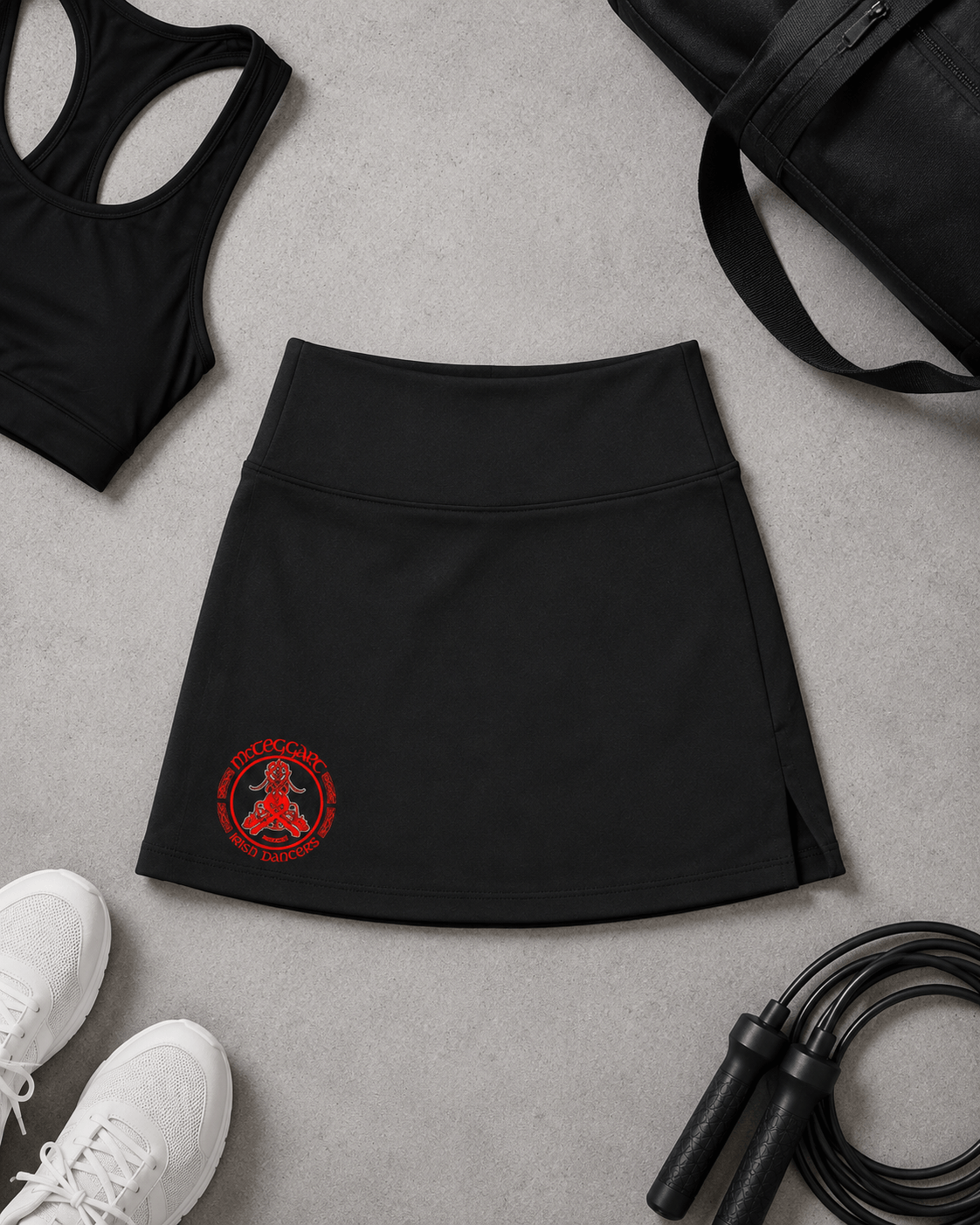 mcteggart irish dancers, A black athletic skirt with a red circular logo on the lower left, styled in a flat lay on a gray textured surface with a black sports bra, black bag, white sneakers, and jump rope.