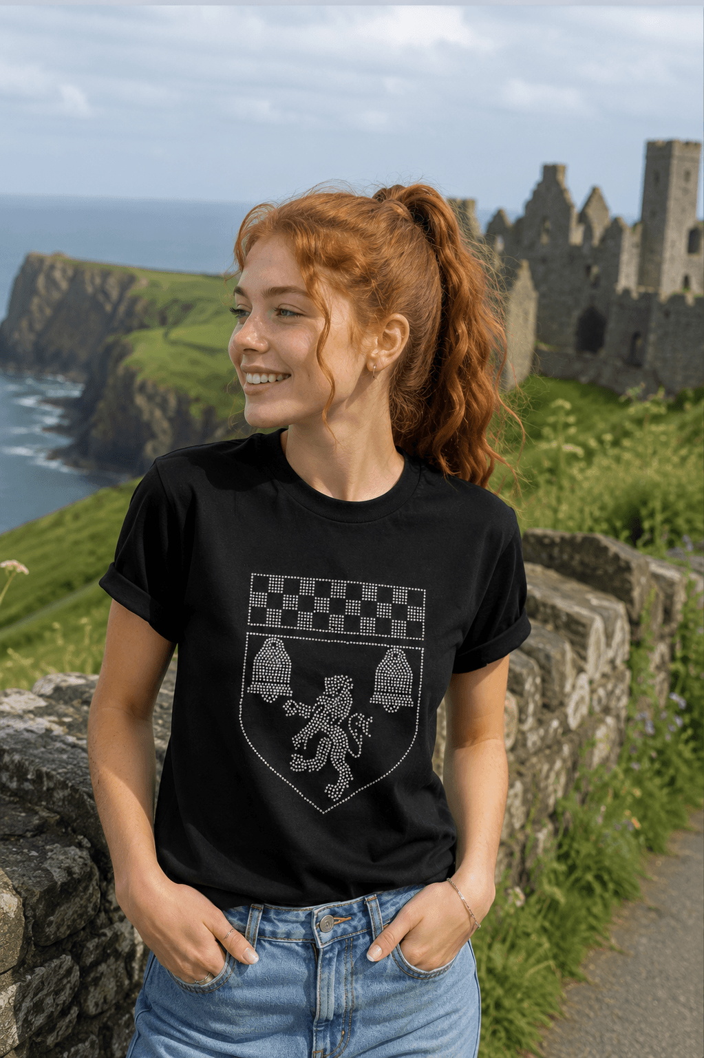 A woman with red hair in a ponytail stands outdoors wearing a black short-sleeve t-shirt with a silver graphic of a shield, bell, and checkerboard pattern. The background shows a scenic coastal landscape with cliffs, sea, and stone ruins.