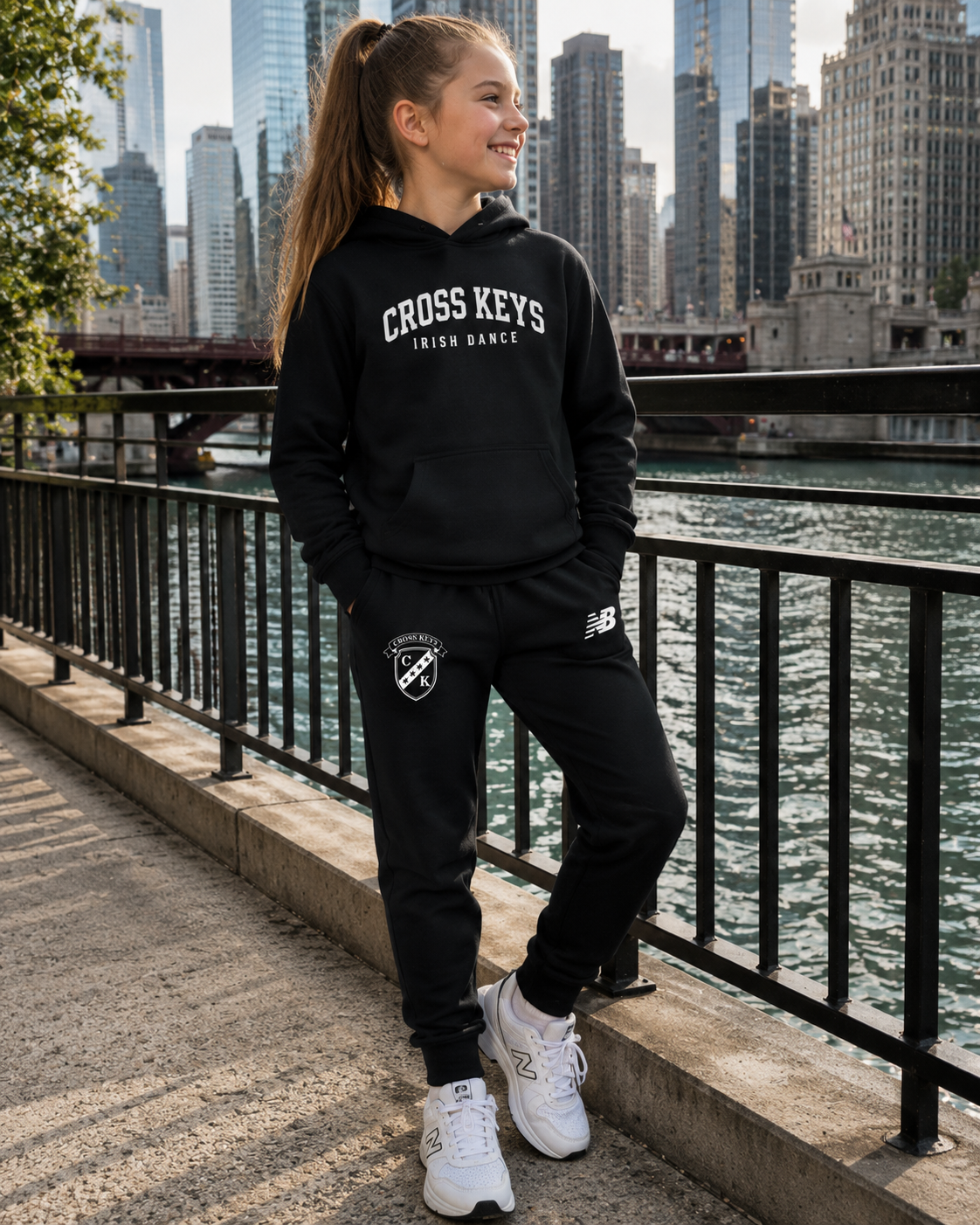 A girl wearing a black Cross Keys Irish Dance hoodie and matching sweatpants stands on a riverside walkway with a city skyline in the background. The hoodie has white text reading 'CROSS KEYS IRISH DANCE' on the chest, and the sweatpants have a small white crest logo near the left thigh.