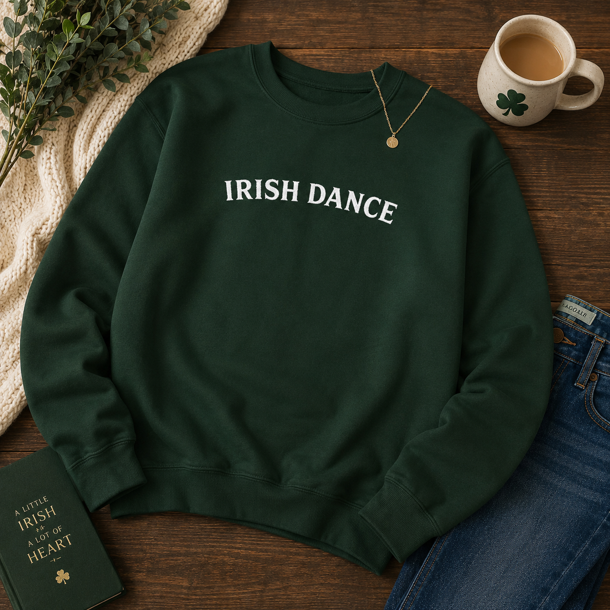 A dark green crewneck sweatshirt with 'IRISH DANCE' printed in white across the chest, styled in a flat lay on a wooden table with a coffee mug, jeans, a book, and a necklace.