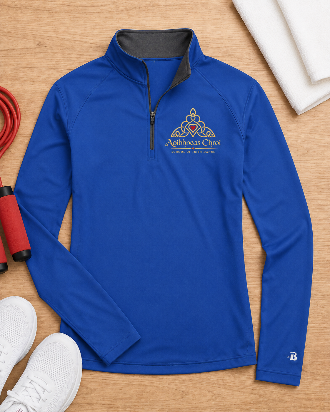 A royal blue long-sleeve quarter-zip athletic top laid flat on a light wood surface, with a gray inner collar, a triangular Celtic knot style chest logo reading 'Aoibhneass Chroi' in gold and white, and a small white logo near the right sleeve cuff. White sneakers, a red jump rope, and folded white towels are placed around it as styling props.