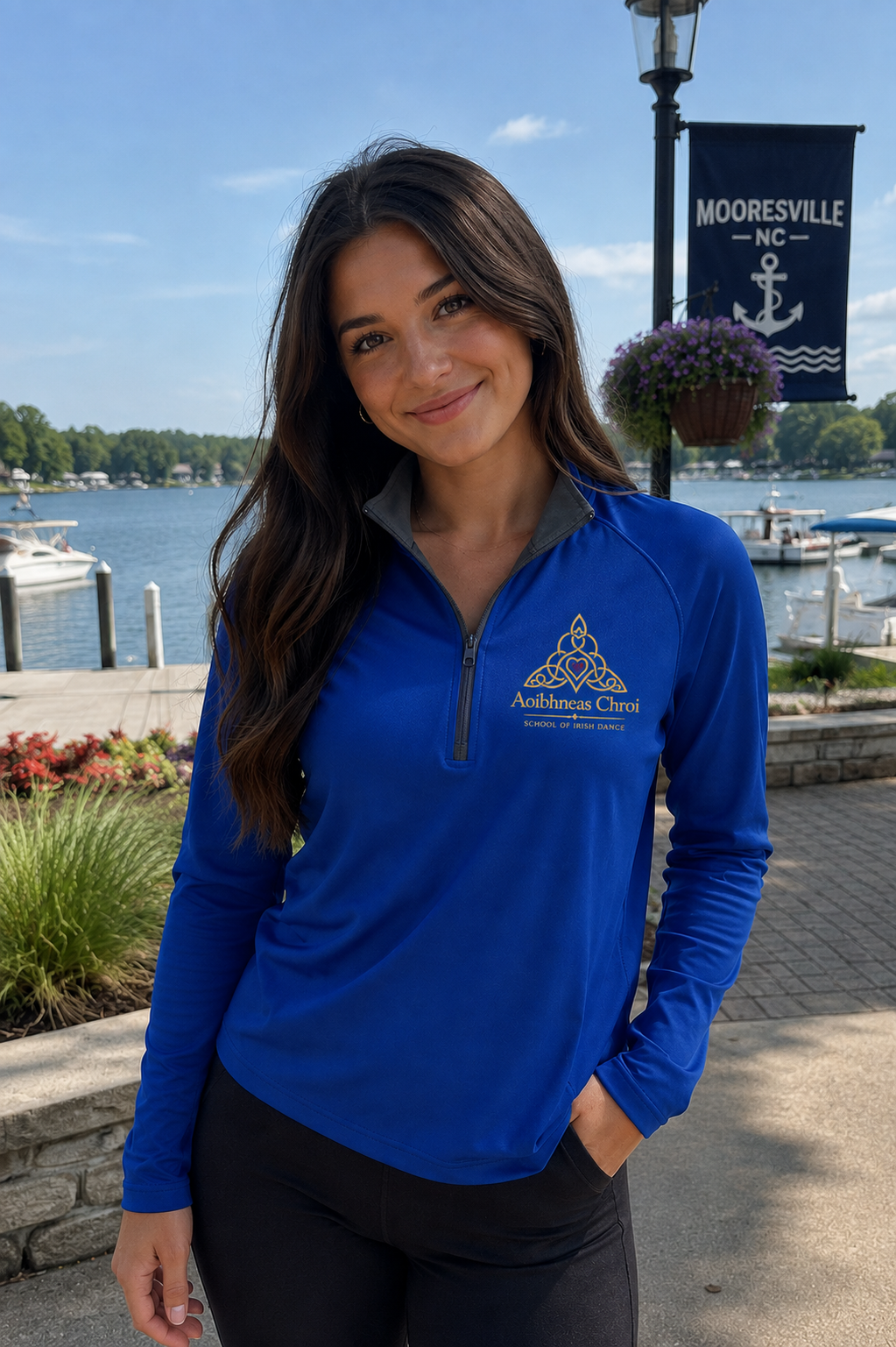 A woman stands outdoors by a waterfront wearing a bright royal blue long-sleeve quarter-zip pullover with a gold and white logo on the left chest. The background shows water, boats, trees, and a black hanging sign that reads Mooresville NC with an anchor.
