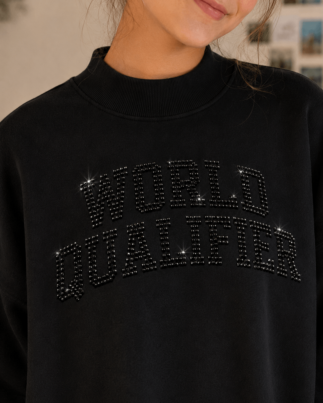 A close-up of a person wearing a black crewneck sweatshirt with 'WORLD QUALIFIER' spelled out in sparkling rhinestone letters across the chest.