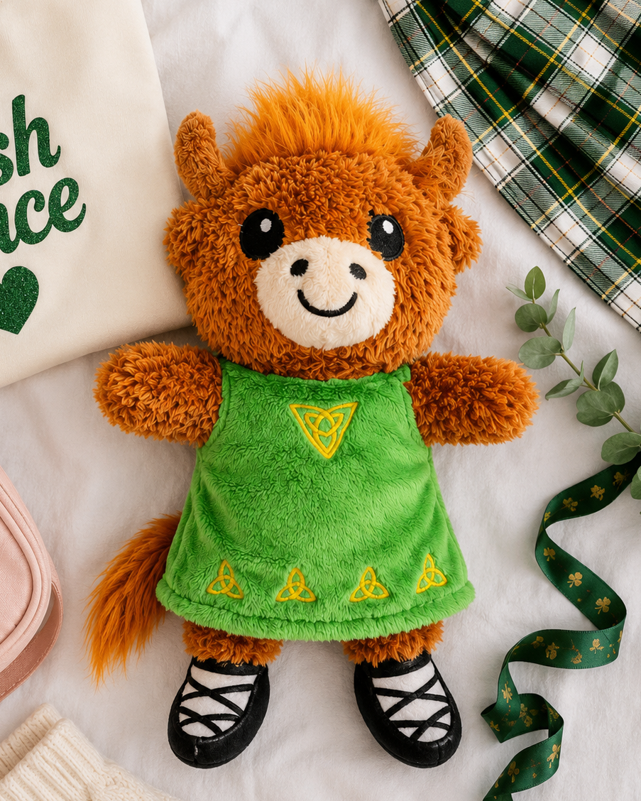 A plush toy horse dressed in a green dress with yellow embroidered triangles and black shoes, styled on a white fabric surface with an Irish-themed shirt, plaid fabric, ribbon, knit socks, and a pink pouch.