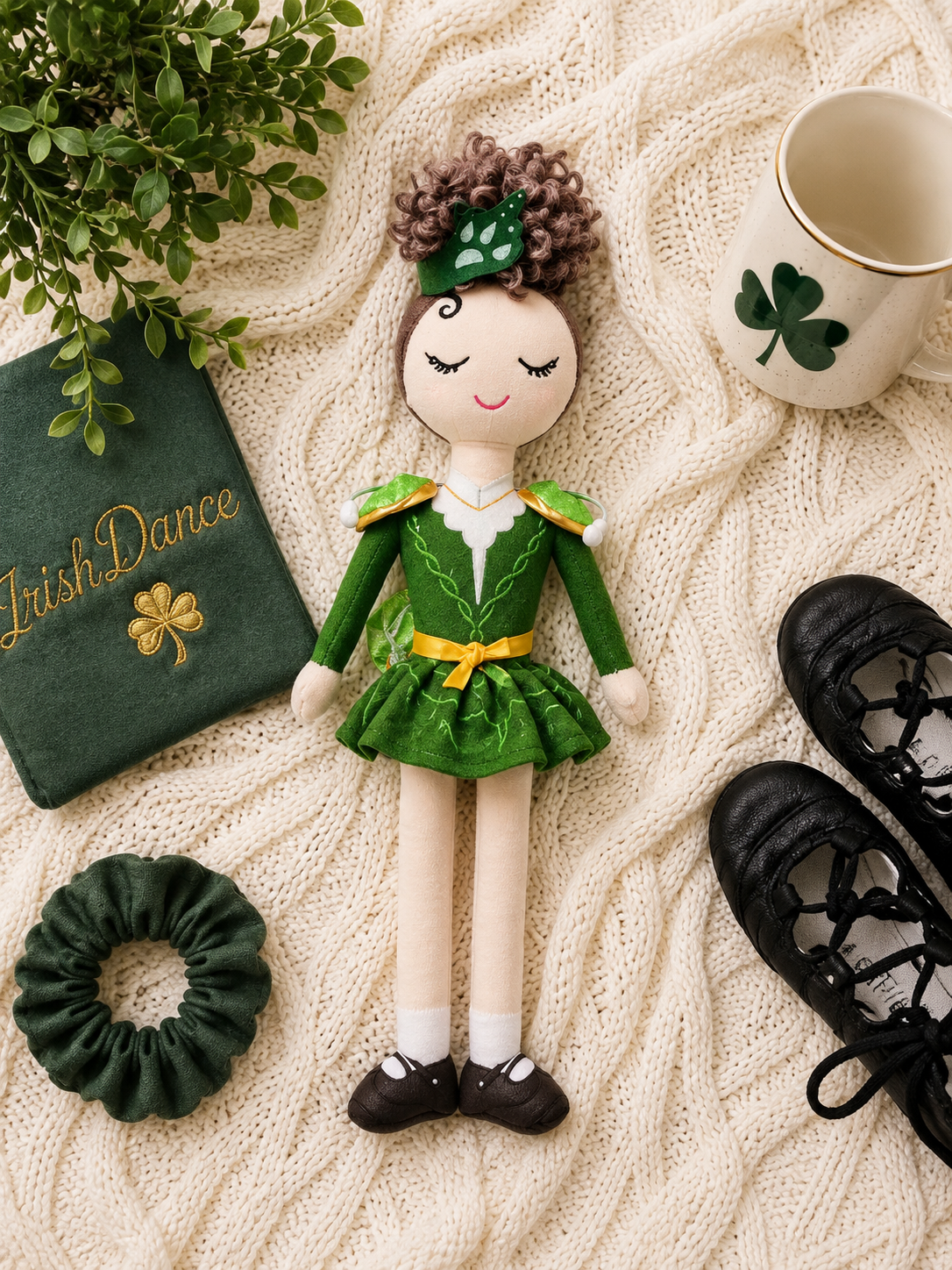 A St. Patrick's Day themed doll dressed in a green leprechaun outfit with a yellow belt, styled on a cream cable-knit blanket with shamrock-themed props including a mug, shoes, scrunchie, book, and plant.