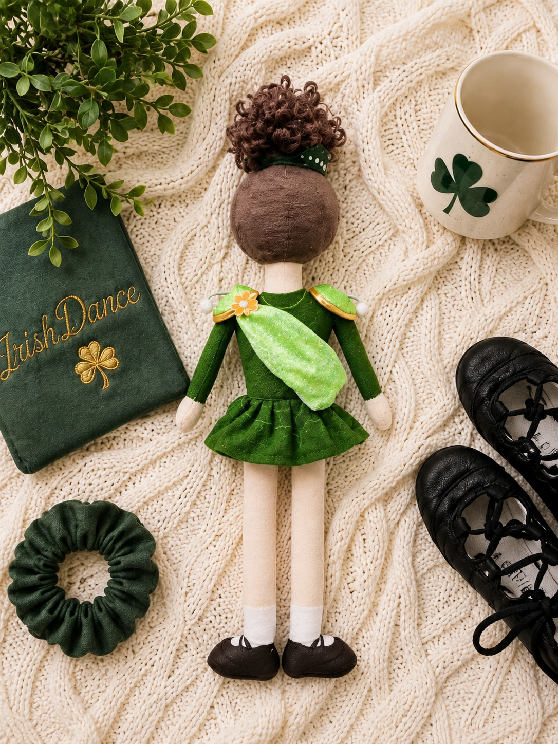 A St. Patrick's Day themed doll dressed in a green dress and scarf, styled in a flat-lay arrangement with a green 'Irish Dance' book, shamrock hair scrunchie, black lace-up shoes, and a cream mug on a beige cable-knit blanket.