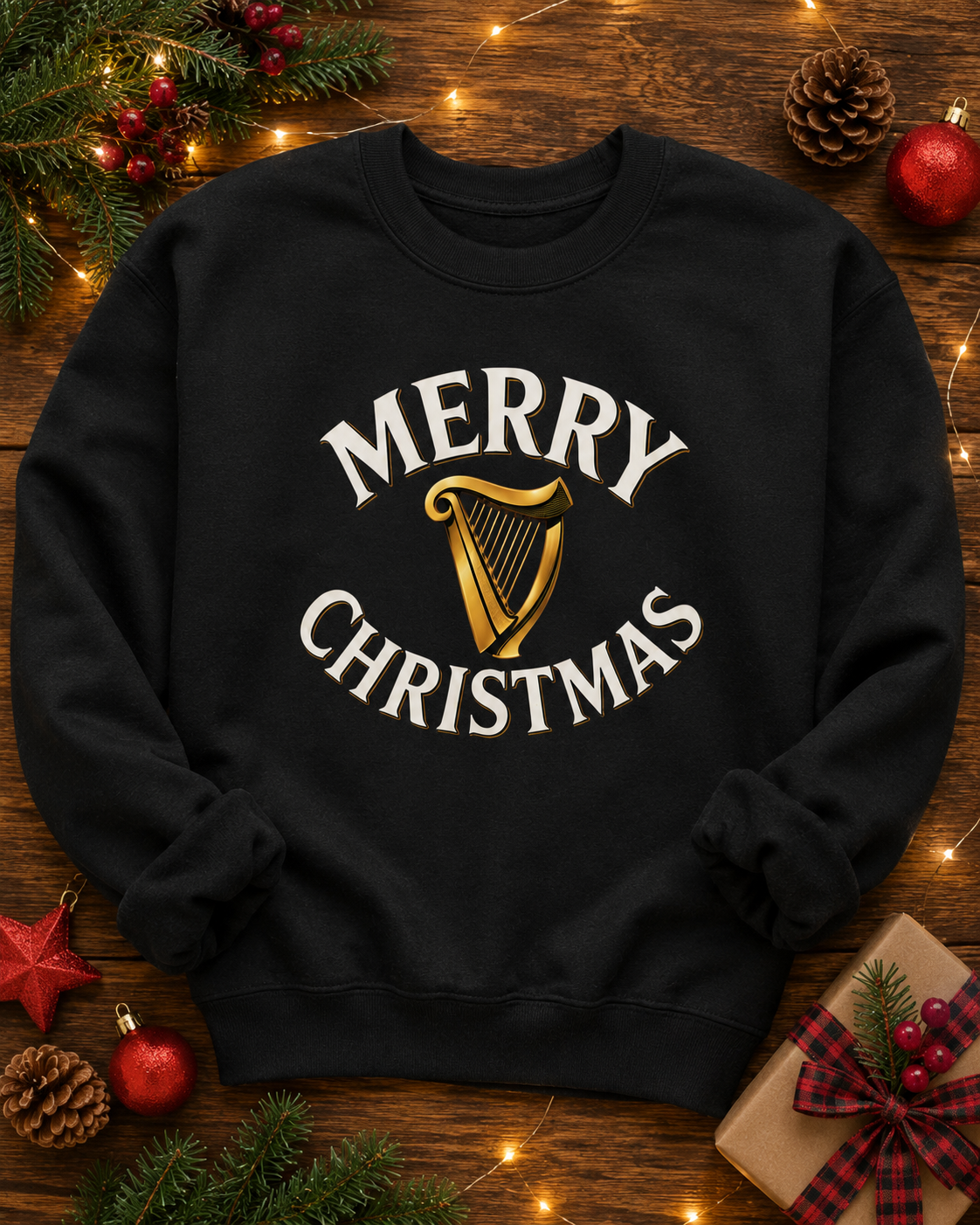 A black crewneck sweatshirt with a 'MERRY CHRISTMAS' graphic featuring a gold harp, laid flat on a rustic wooden background decorated with pine branches, lights, ornaments, and a wrapped gift.