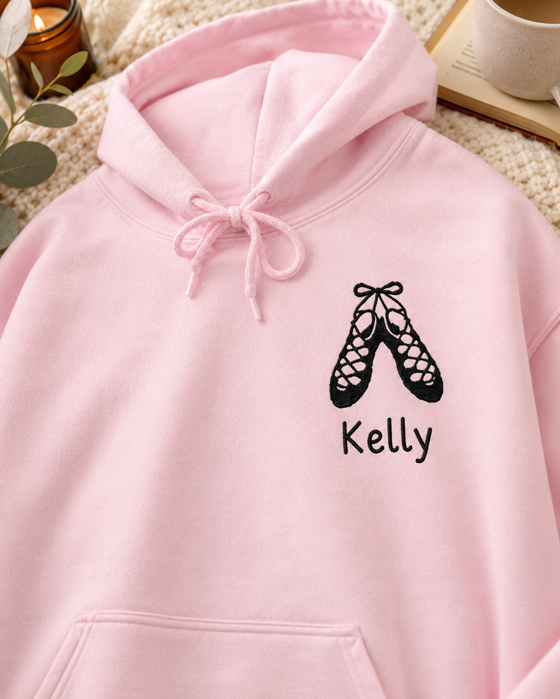 A light pink hoodie laid flat with a black ballet shoe graphic and the name 'Kelly' printed on the chest, styled with a knit blanket, candle, and book in the background.