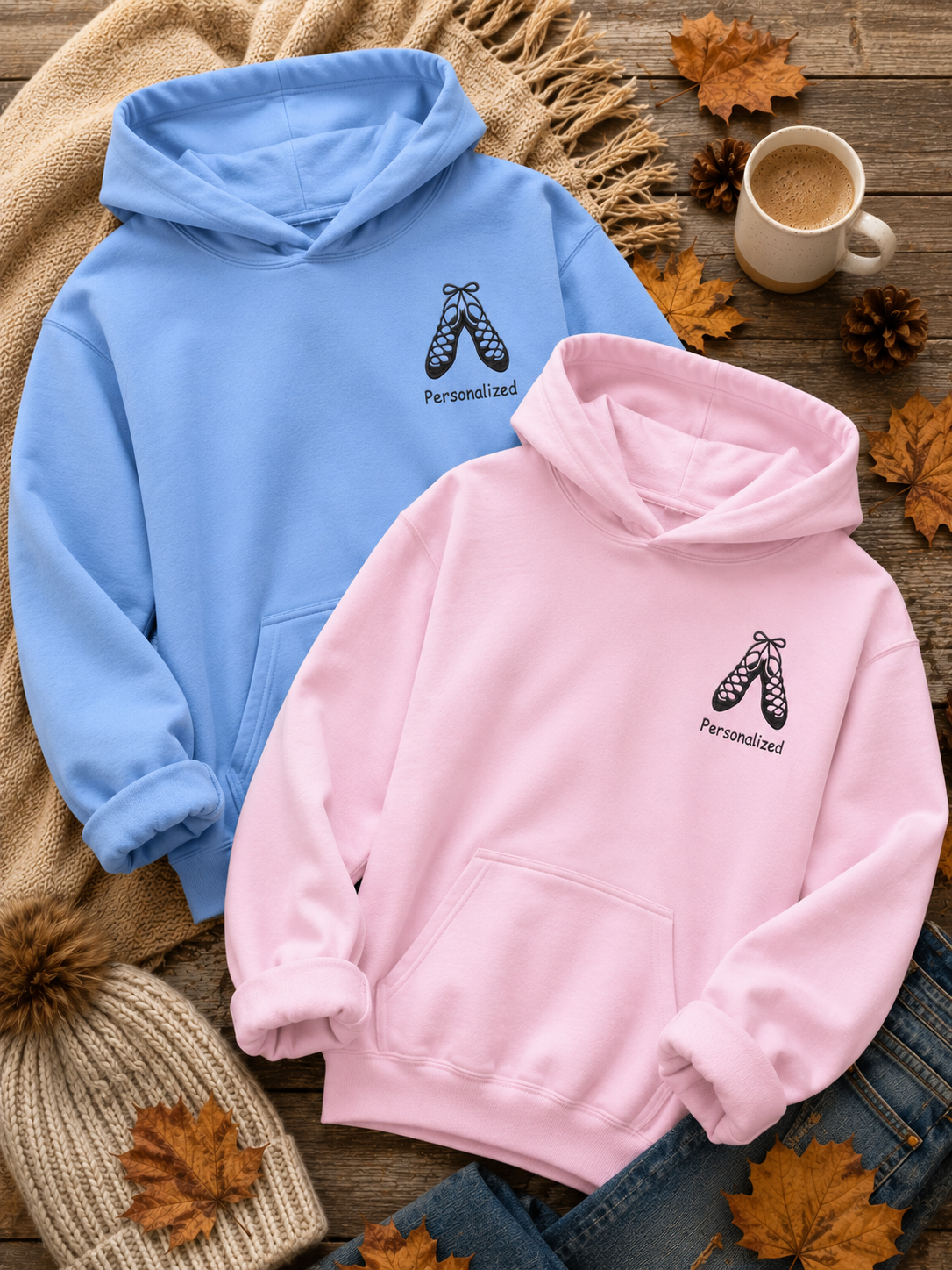 Two personalized pullover hoodies, one light blue and one light pink, laid flat on a rustic wooden surface with autumn props including a knit beanie, a cup of coffee, pine cones, and fallen leaves. Each hoodie features a black graphic of ballet slippers with the word 'Personalized' underneath.