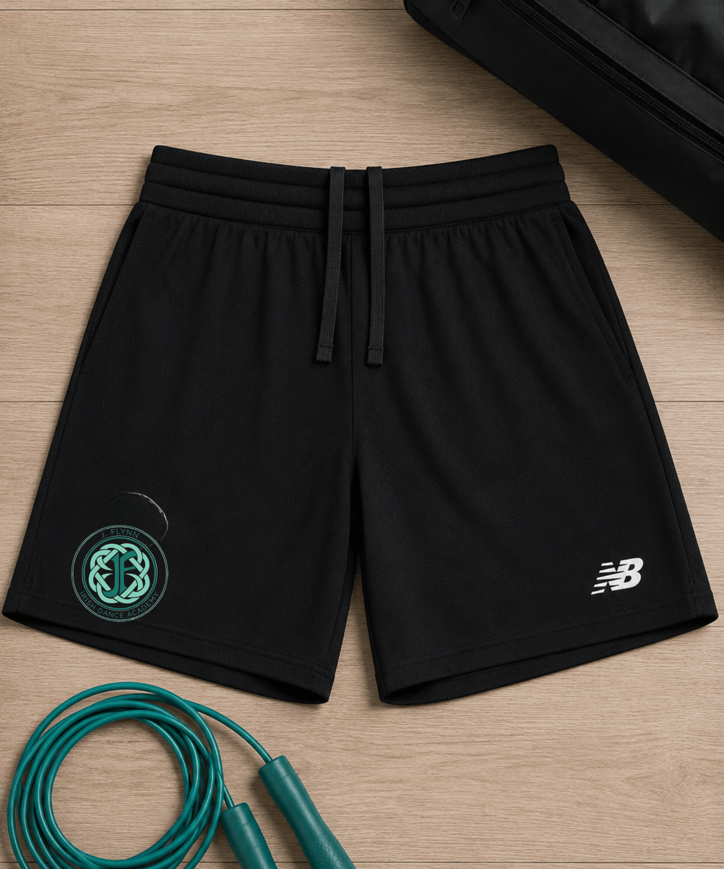 Black athletic shorts with a white New Balance logo on the left leg and a green circular logo on the right leg, laid flat on a light wood background with a jump rope and a black bag as props.