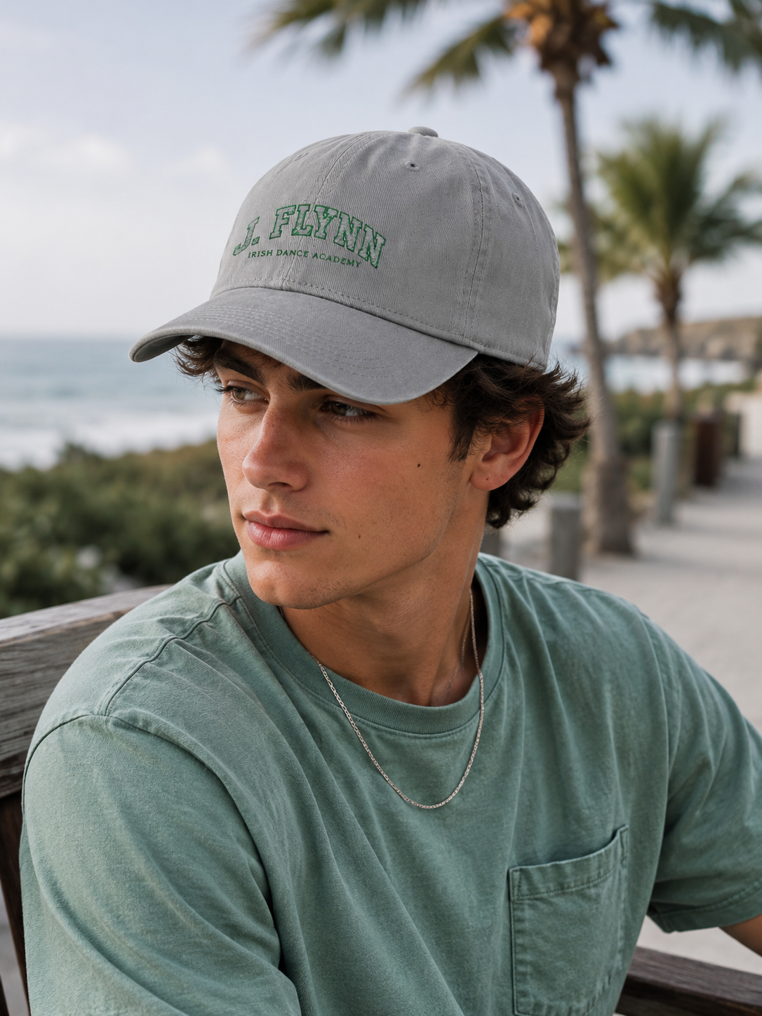 A man wearing a light gray baseball cap with green embroidered text reading 'J. FLYNN' and a matching sage green t-shirt, photographed outdoors on a beachside walkway with palm trees and the ocean in the background.
