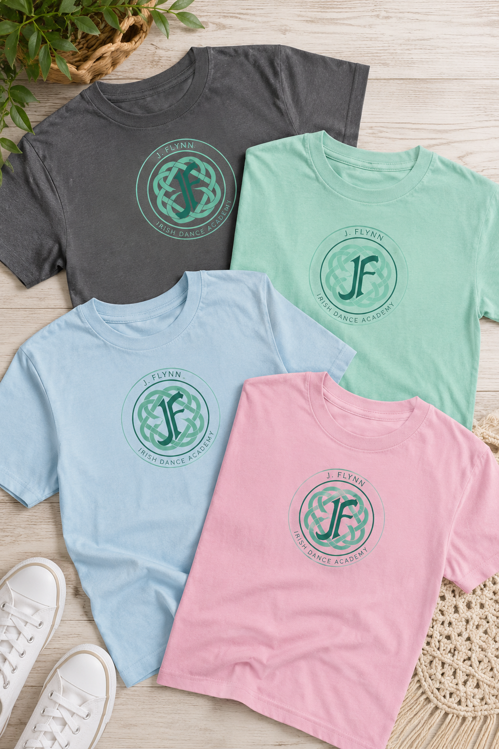 Four short-sleeve crewneck T-shirts in dark gray, mint green, light blue, and pink, arranged in a flat lay on a light wood surface. Each shirt features a circular teal graphic with a stylized 'JF' monogram and the words 'J.