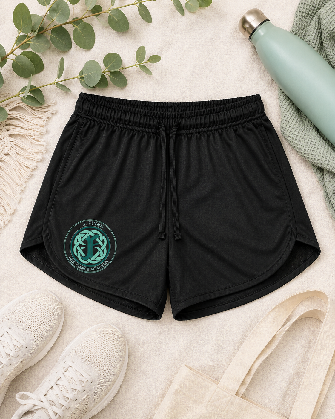 Black athletic shorts with a drawstring waist and a teal circular logo on the left leg, styled in a flat lay with white sneakers, a canvas tote bag, a mint green water bottle, and eucalyptus branches on a cream fabric background.