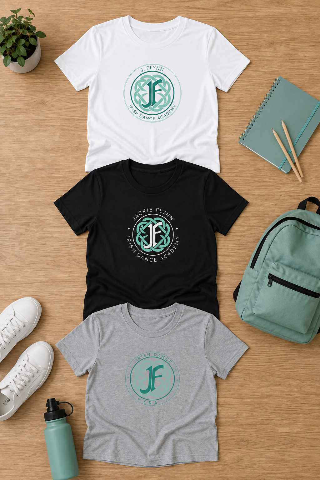 Three short-sleeve graphic T-shirts in white, black, and heather gray are arranged in a flat lay on a light wood surface, surrounded by a plant, notebook, pens, shoes, water bottle, and backpack. Each shirt features a circular teal logo with a stylized letter F and the words 'FJLN FLYING JUNGLE BALANCE ACTOR' or similar themed text.