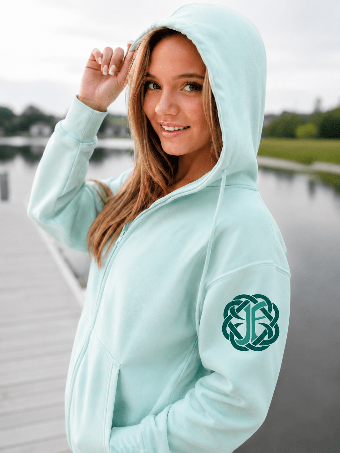 A woman wearing a mint green hooded sweatshirt with a dark green Celtic knot monogram on the left sleeve, standing on a wooden dock by a calm lake.