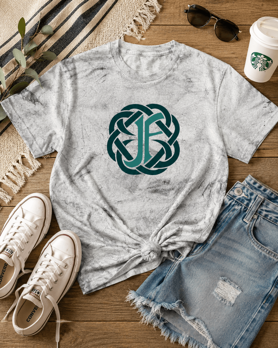 A light gray acid-wash t-shirt with a teal Celtic knot monogram graphic, styled in a flat lay with denim shorts, white sneakers, sunglasses, a Starbucks cup, and a striped blanket on a wooden floor.