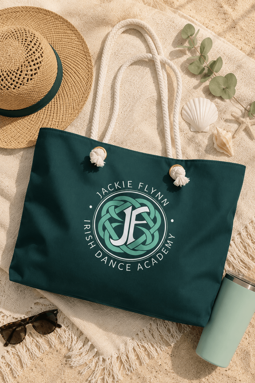 A dark teal tote bag with white rope handles and a circular 'Jackie Flynn Irish Dance Academy' logo printed on the front, styled on a beige blanket with a straw hat, sunglasses, a tumbler, and seashells.