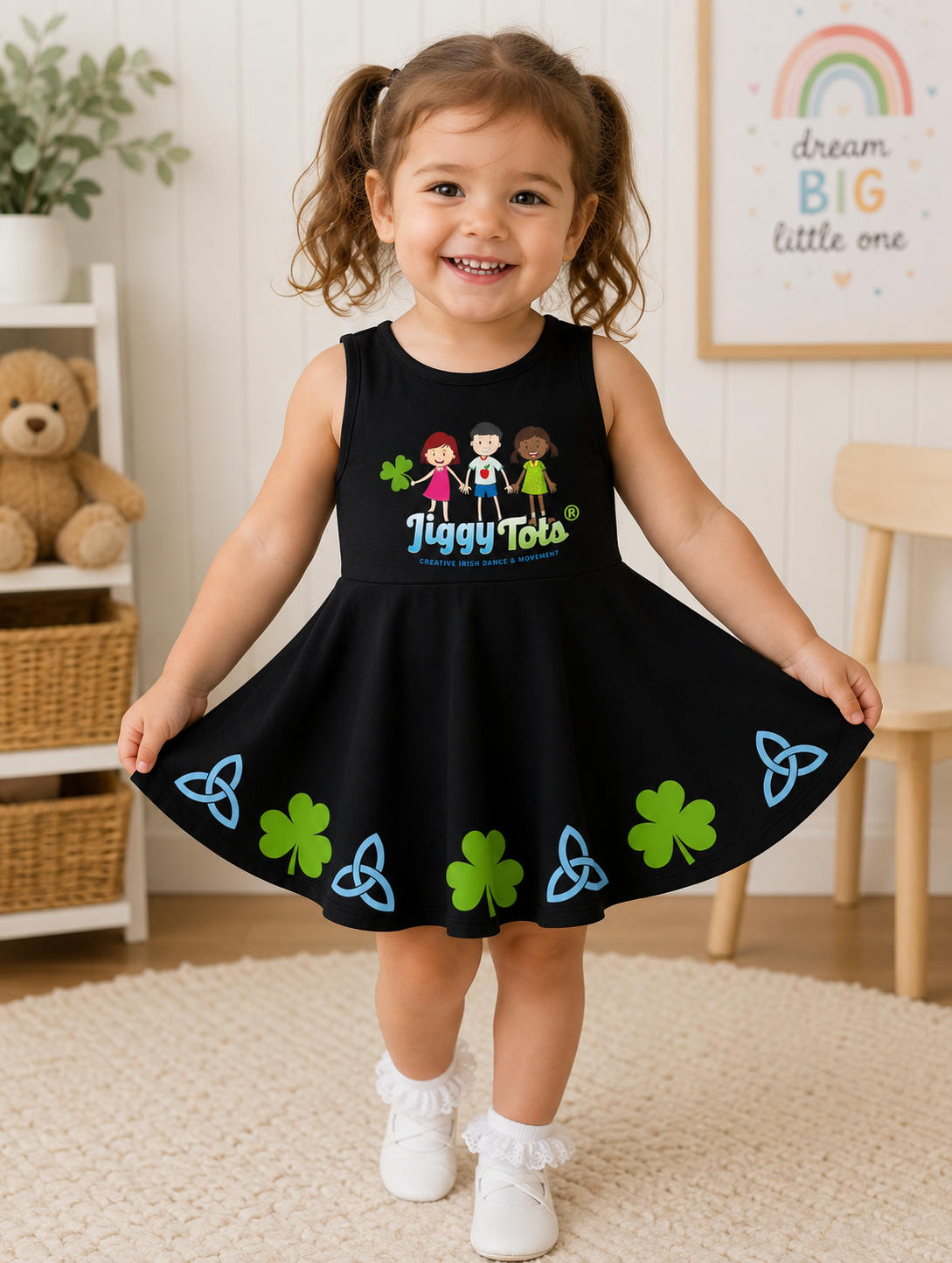 A young girl with brown pigtails stands in a child's room wearing a black sleeveless dress with a colorful 'Jiggy Tots' graphic on the chest and green shamrock and blue Celtic knot patterns along the skirt.