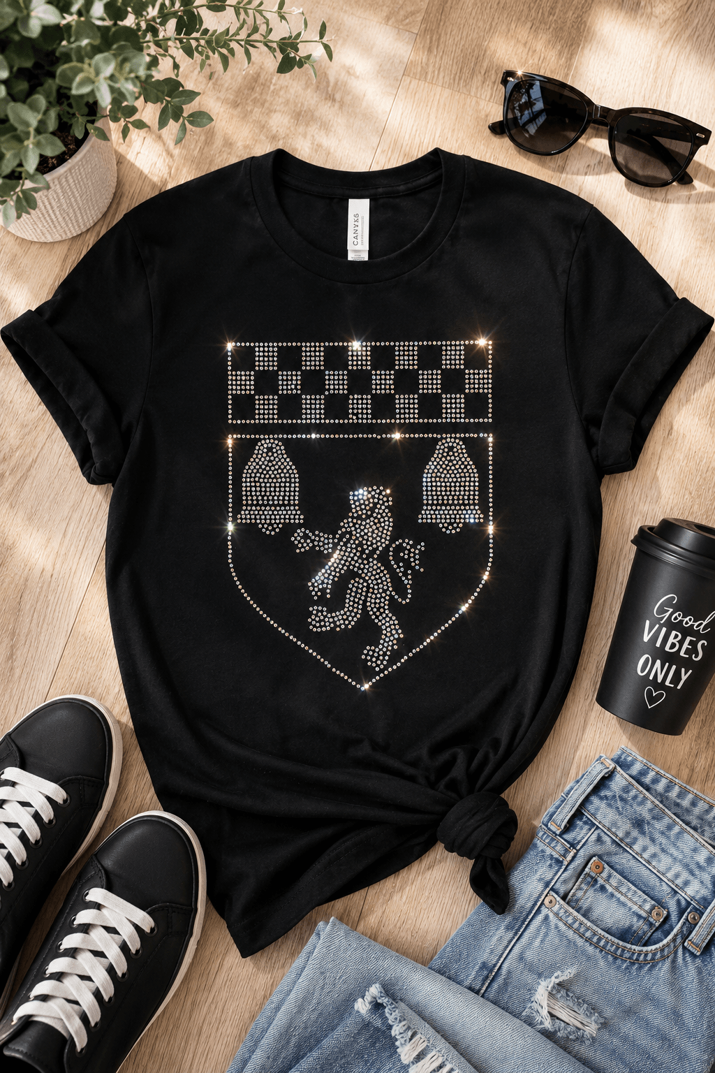 A black short-sleeve T-shirt laid flat on a light wood floor, styled with jeans, black sneakers, sunglasses, a potted plant, and a black coffee cup. The shirt has a large rhinestone-style crest graphic on the front featuring a lion and checkerboard details.