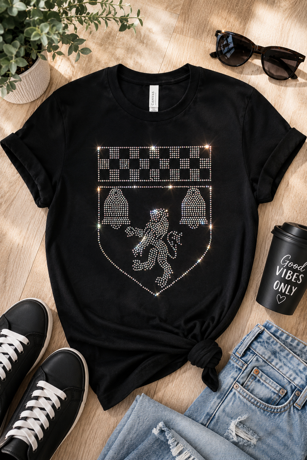 A black short-sleeve T-shirt laid flat on a light wood floor, styled with jeans, black sneakers, sunglasses, a potted plant, and a black coffee cup. The shirt has a large rhinestone-style crest graphic on the front featuring a lion and checkerboard details.