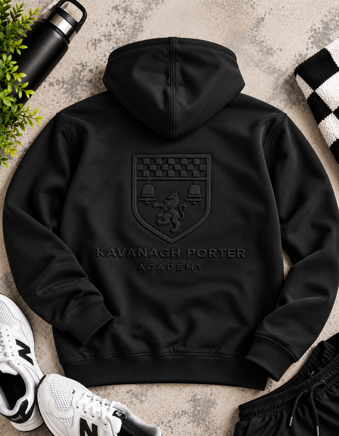 A black hooded sweatshirt laid flat on a textured stone surface, showing the back with an embossed Kavanagh Porter Academy crest and text. White sneakers, a black water bottle, greenery, and a black-and-white checkered cloth are placed around it as styling props.