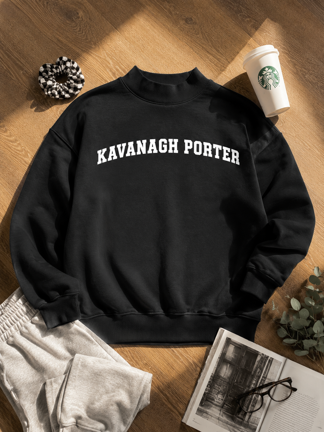 A black crewneck sweatshirt with 'KAVANAGH PORTER' printed in white arched text across the chest, styled in a flat lay on a wooden floor with a white Starbucks cup, gray sweatpants, a black-and-white scrunchie, glasses, and an open book.
