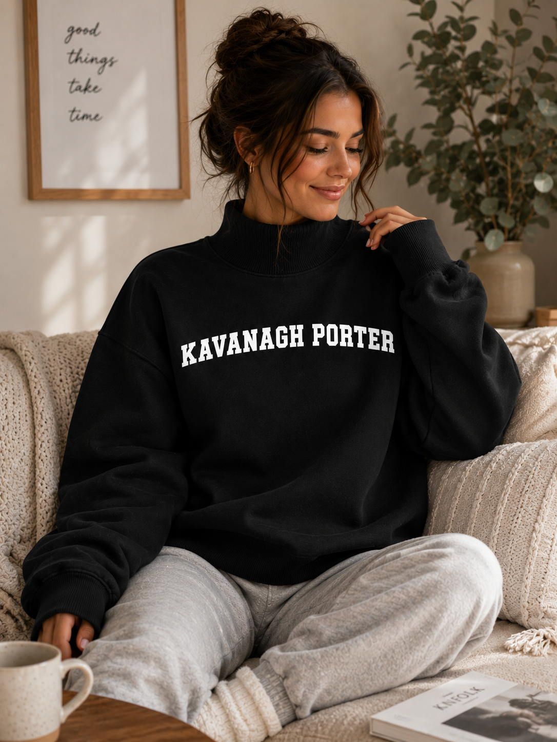 A woman sitting on a cozy knit couch wearing a black sweatshirt with 'KAVANAGH PORTER' printed in white across the chest, paired with light gray sweatpants. A mug and a book are on the table in front of her, with a framed print and a potted plant in the background.