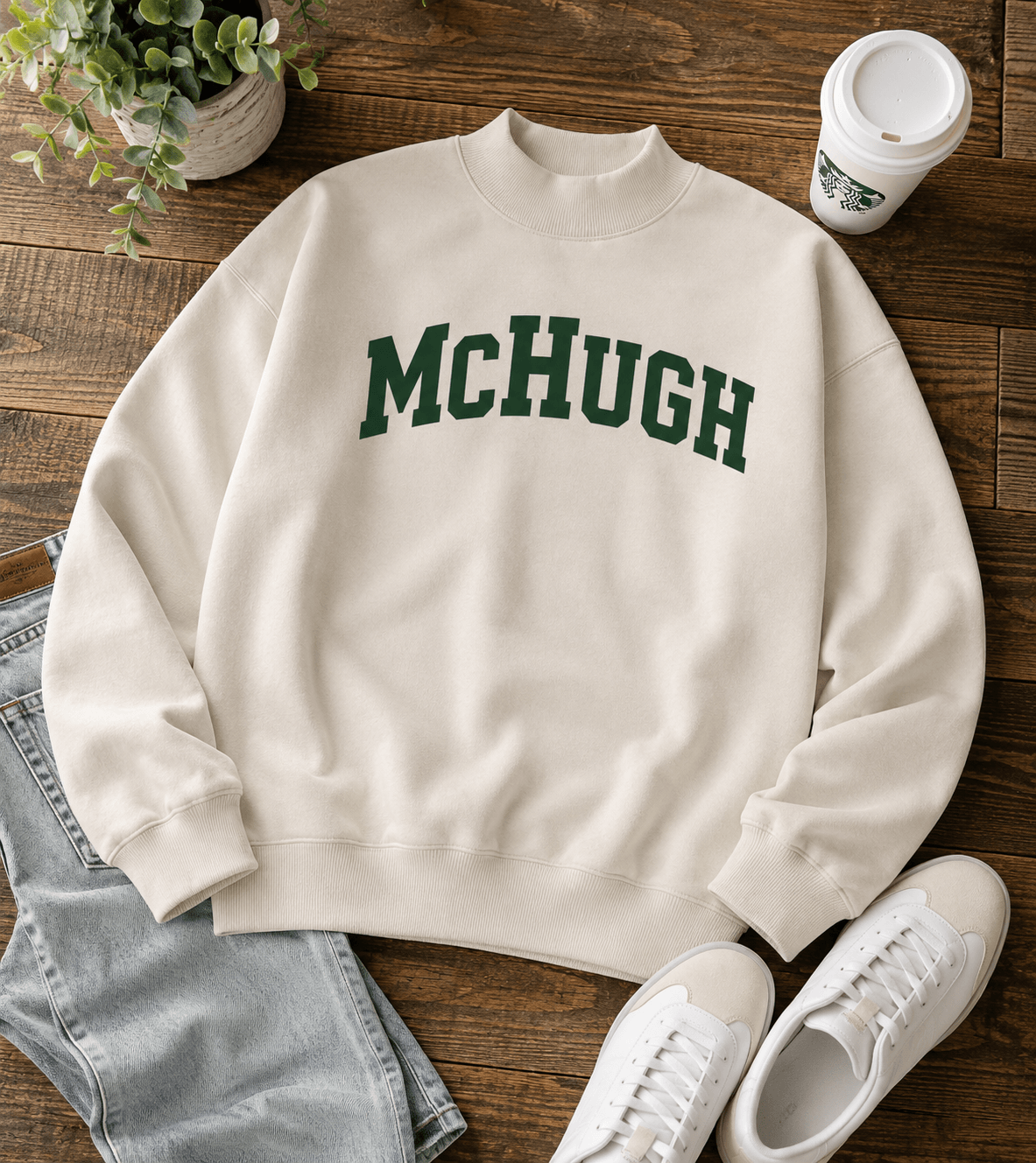 An off-white mock neck sweatshirt with 'MCHUGH' printed in dark green varsity letters across the chest, styled in a flat-lay arrangement on a wooden table with jeans, white sneakers, a coffee cup, and a potted plant.