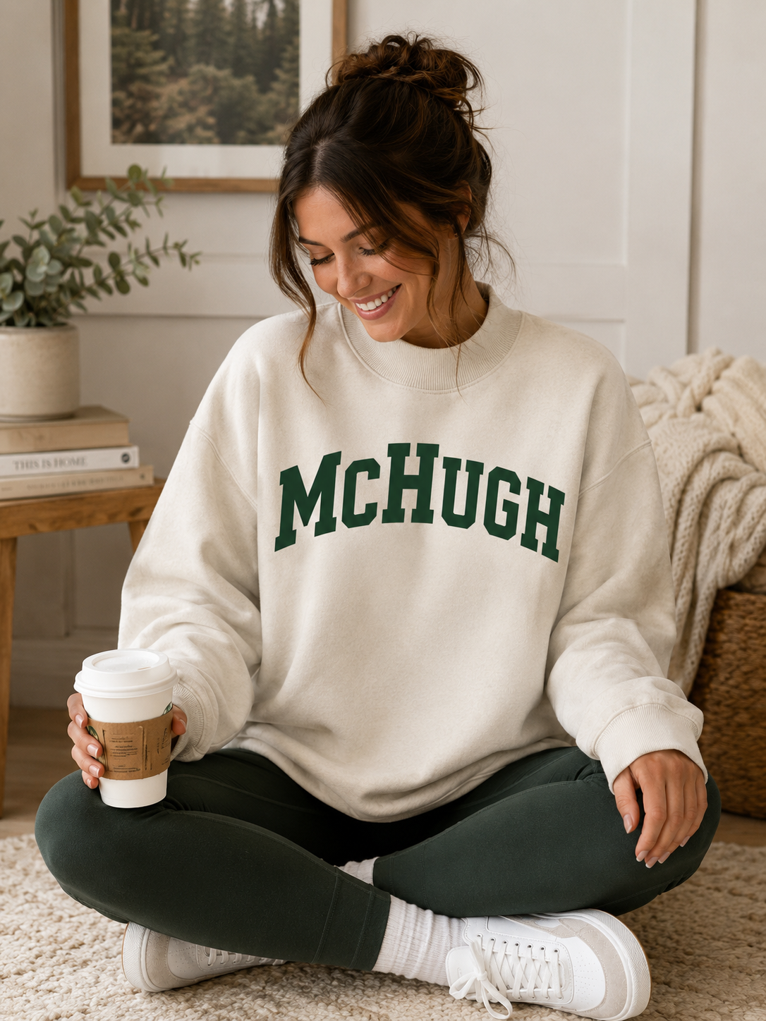 A woman sitting cross-legged on a beige carpet in a cozy room, wearing a cream sweatshirt with 'McHUGH' printed in dark green across the chest, dark green leggings, white socks, and white sneakers, while holding a takeaway coffee cup.
