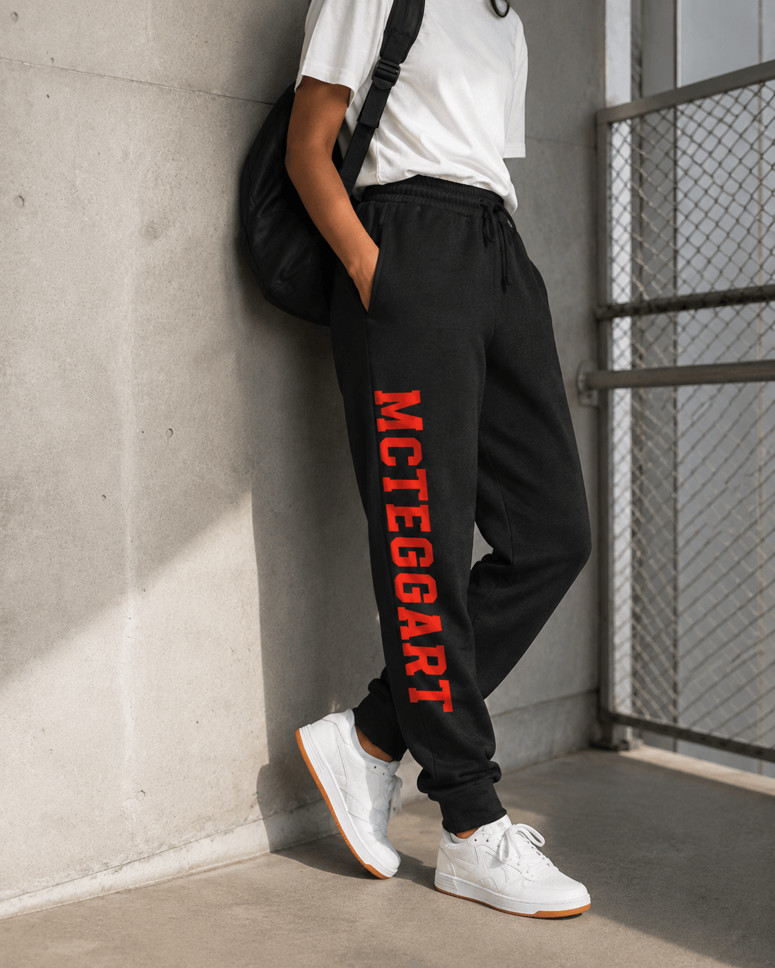 mcteggart irish dancers, A person wearing black sweatpants with bold red vertical 'MCteggart' text down one leg, paired with a white t-shirt and white sneakers, leaning against a concrete wall.